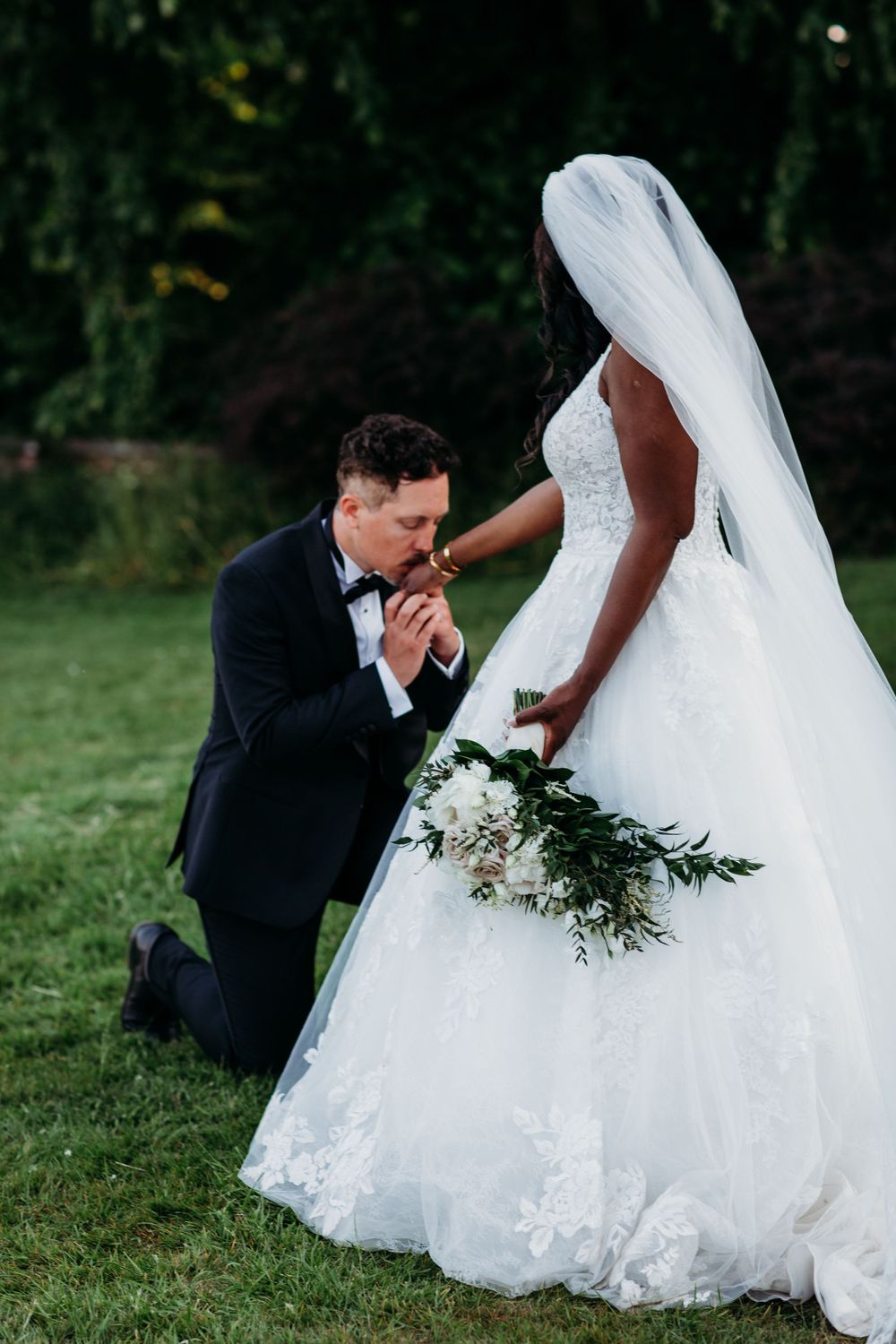 Tender moment captured as groom kisses bride's hand outdoors with flowing white gown and floral bouquet.