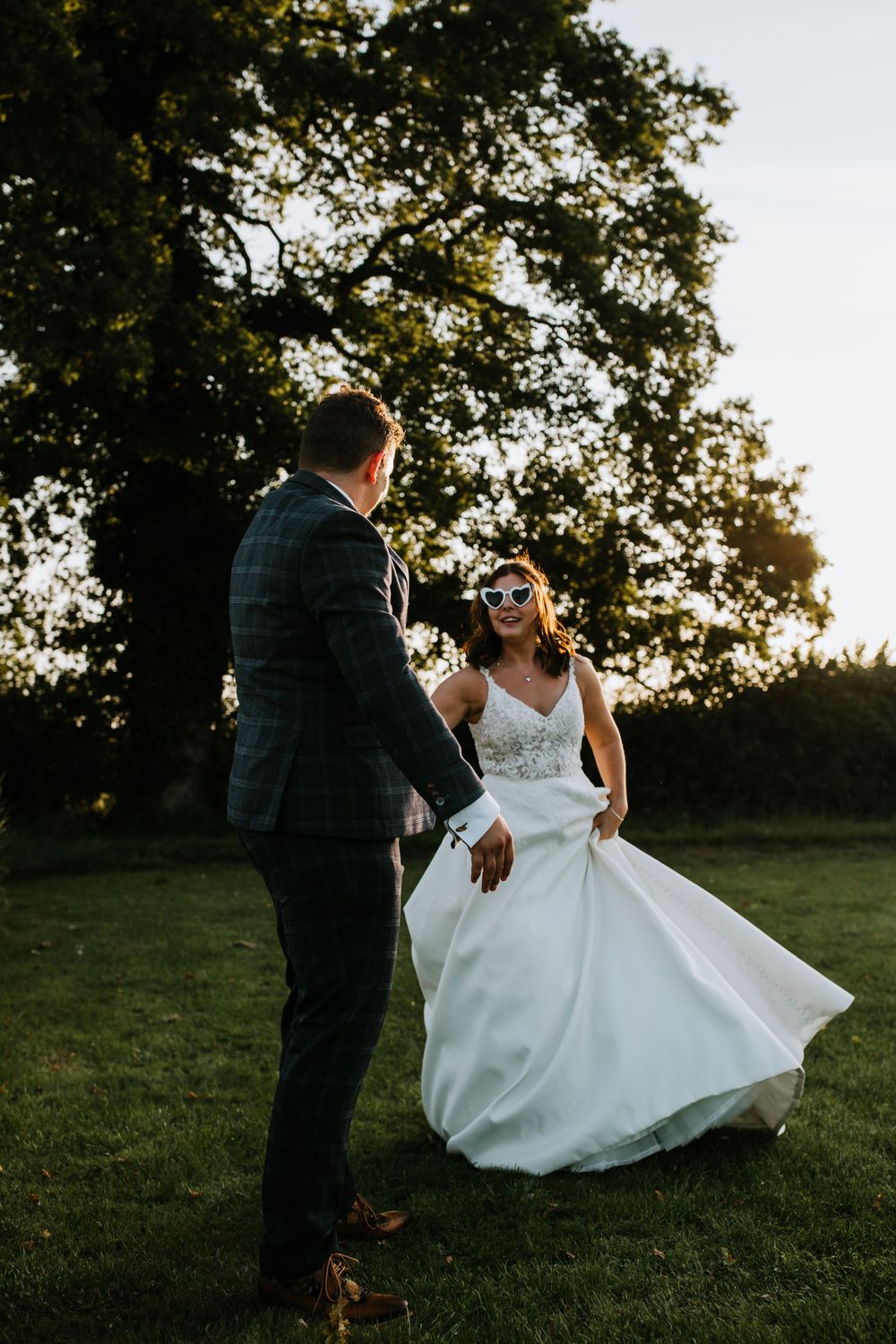Wedding couple dances playfully on grass during golden hour outdoor celebration.