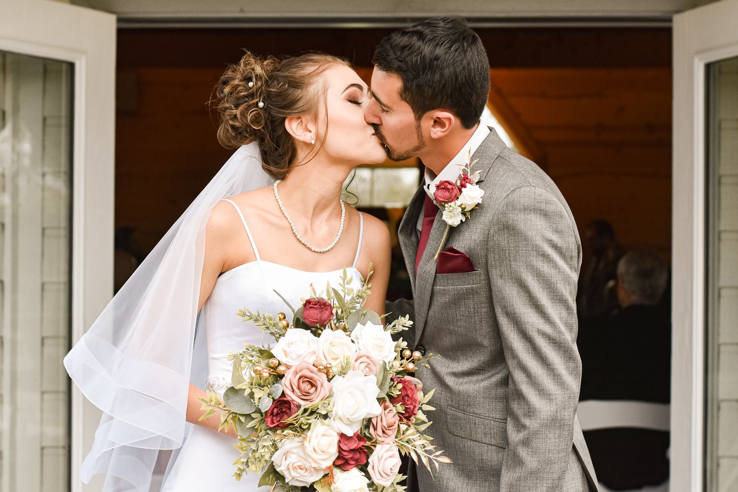 Happy newlyweds sharing a kiss while bride holds beautiful pink and burgundy bouquet.
