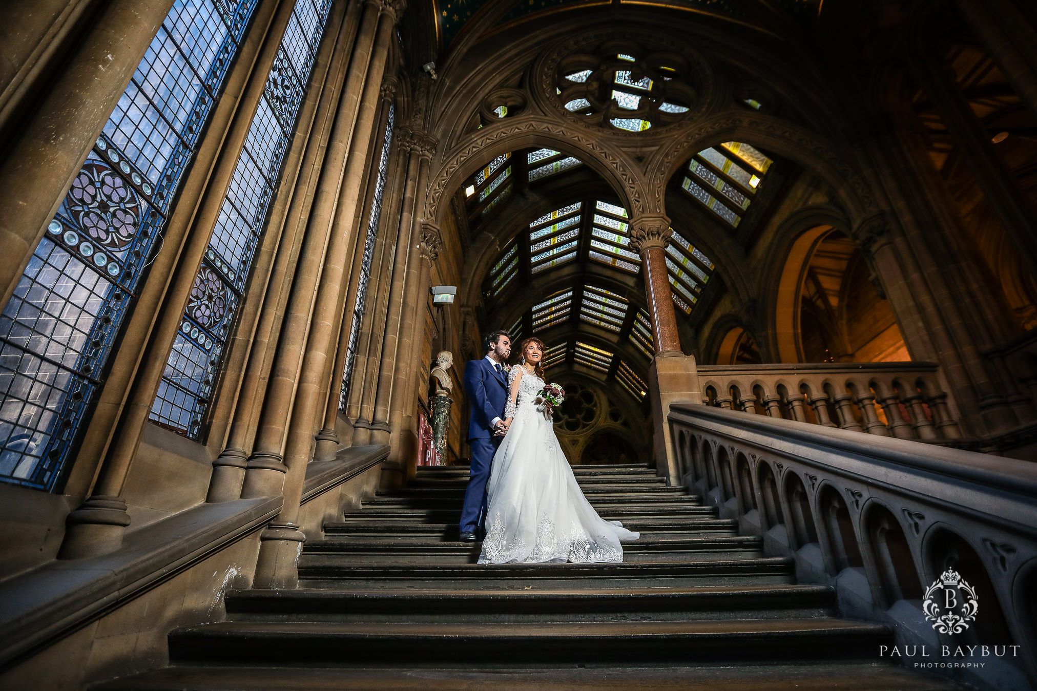 A dramatic wedding photo on grand stone stairs with ornate gothic architecture and stained glass windows.