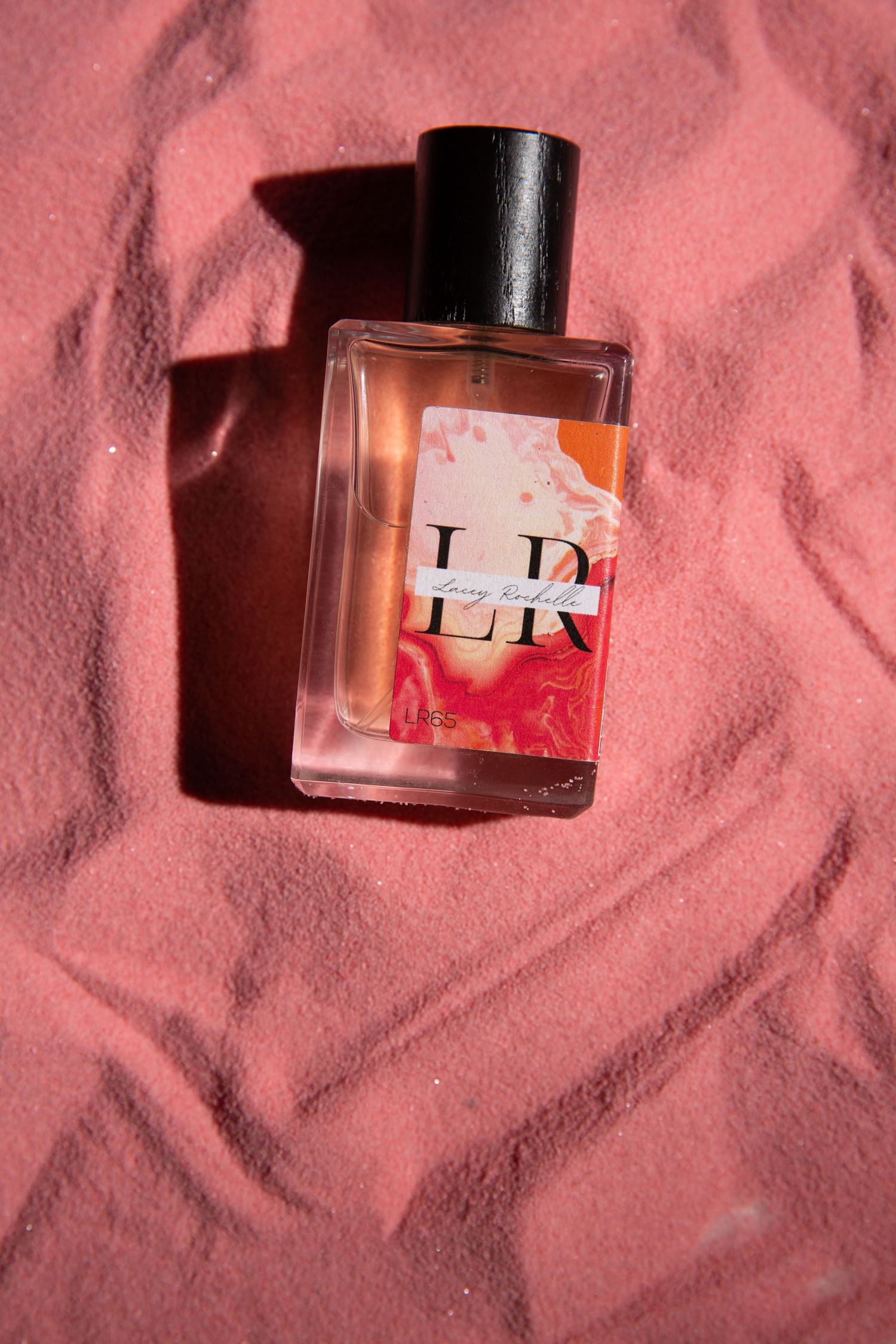 A glass perfume bottle with 'LR' branding sits on a pink textured fabric background casting dramatic shadows.