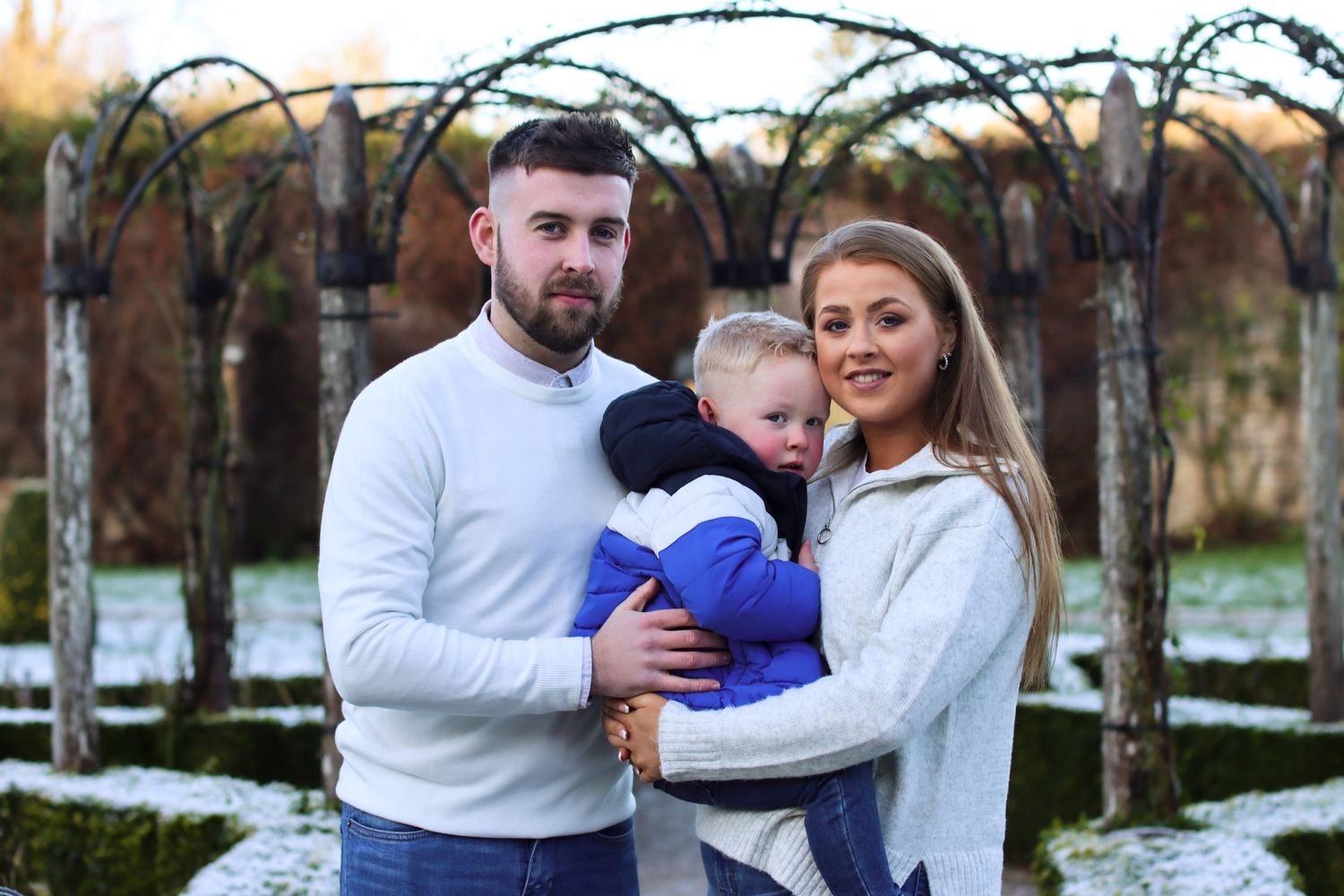 A family portrait outdoors with a small baby in winter clothing standing in front of a garden arch.