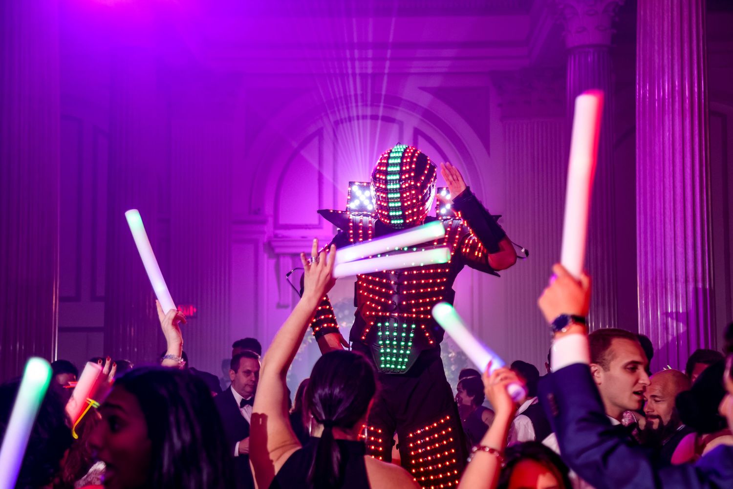 Party guests dance under purple lighting with LED sticks raised in celebration.