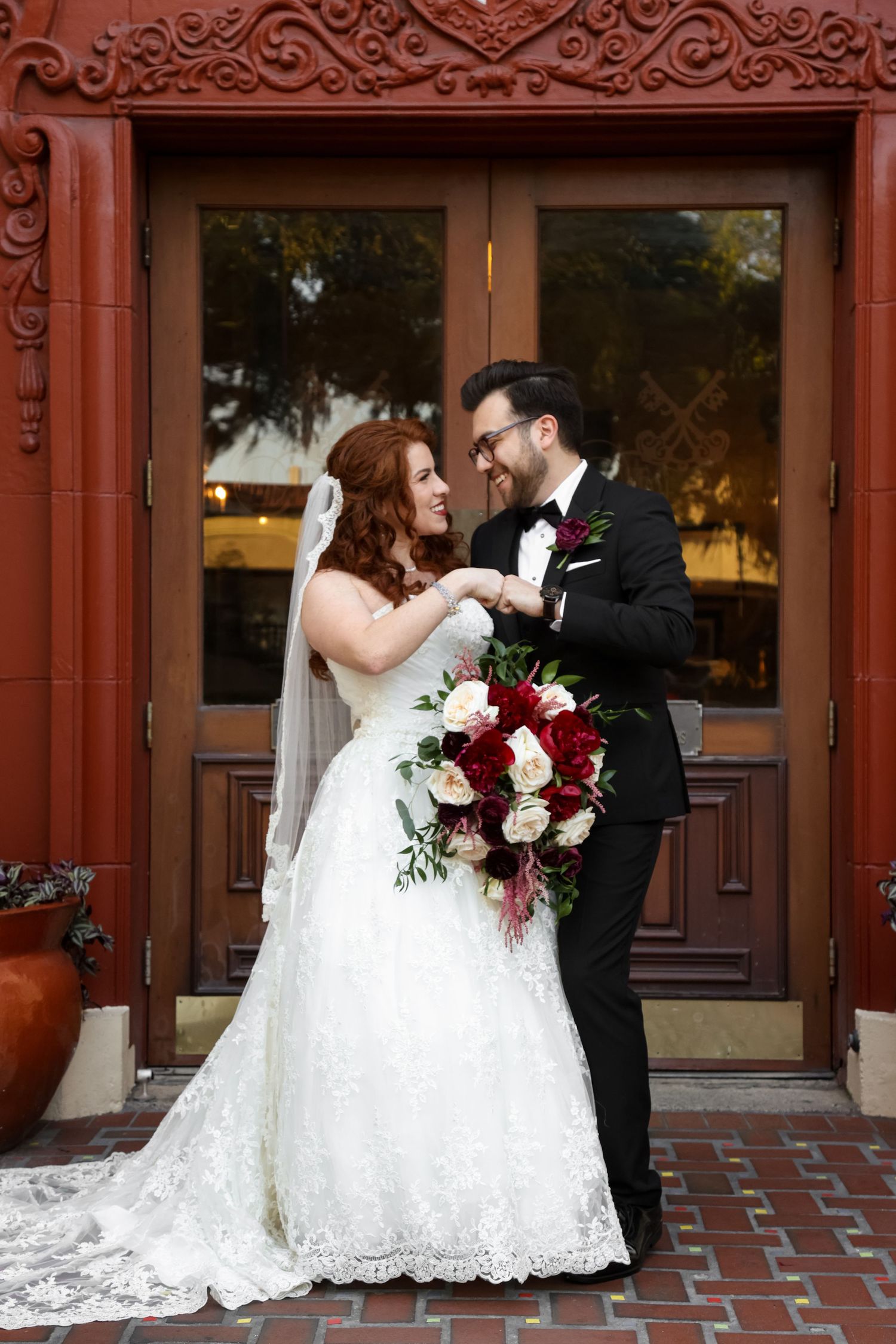A romantic wedding moment between newlyweds in front of an ornate red door with a burgundy and white floral bouquet.
