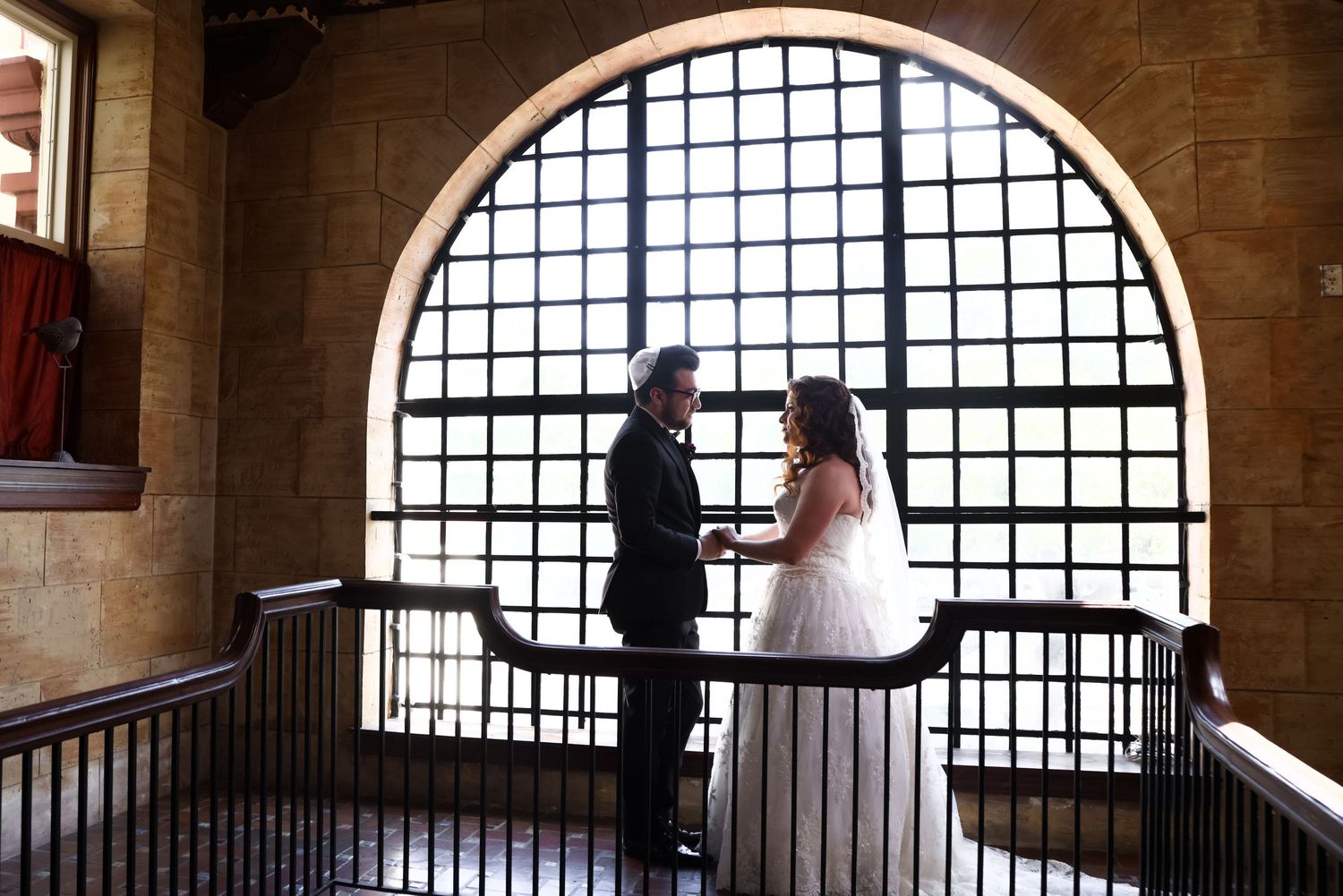 A romantic couple stands in front of a grand arched window with gridded panes inside a stone building interior.