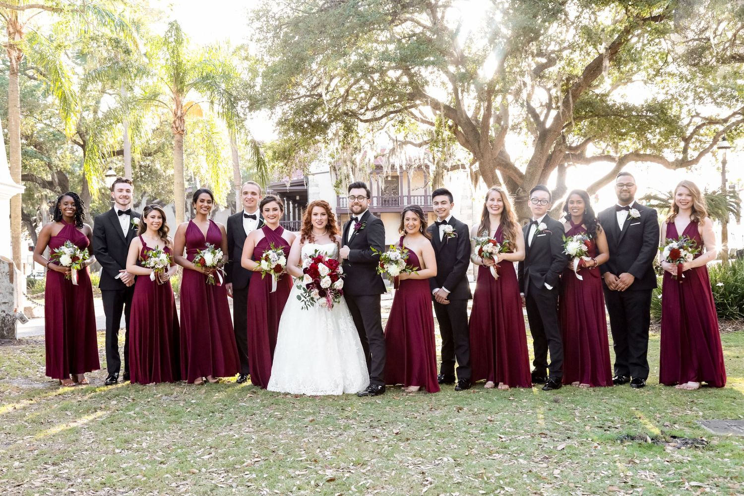 A wedding party poses outdoors in the plaza in ST Augustine with burgundy bridesmaid dresses and formal attire under large shady trees.