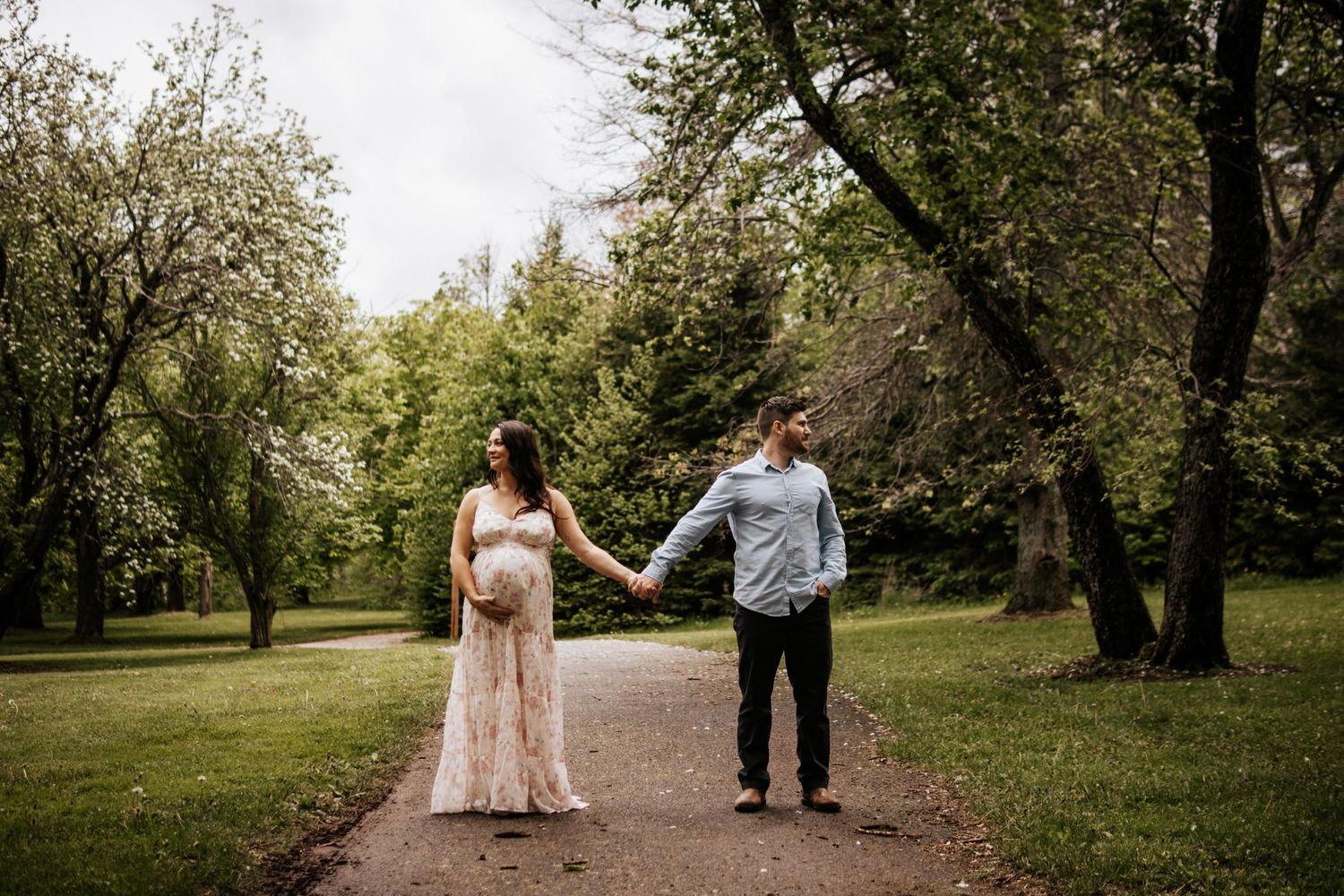 A couple holds hands while walking down a tree-lined path in a lush green park.