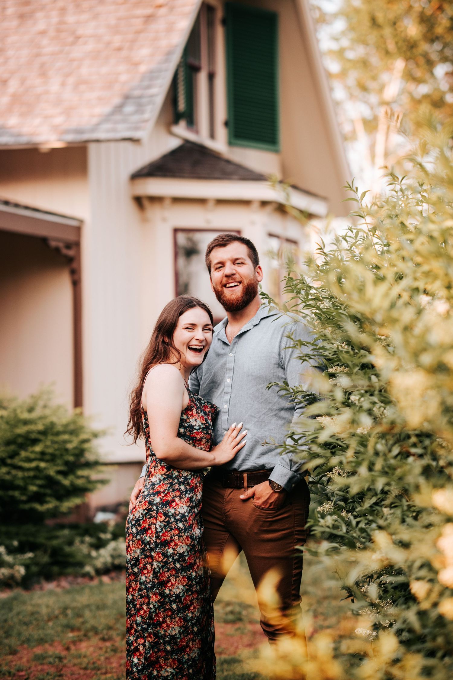 A joyful couple embraces and laughs together in front of a white house with green shutters.