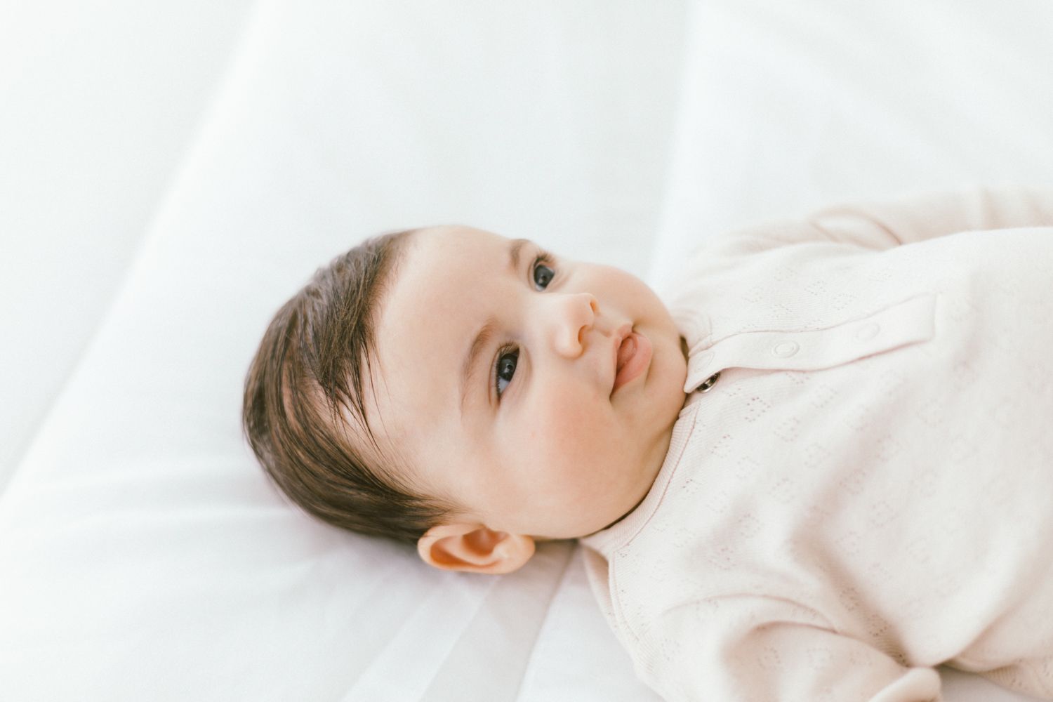 Baby in beige outfit lying on white bedding looking at camera.
