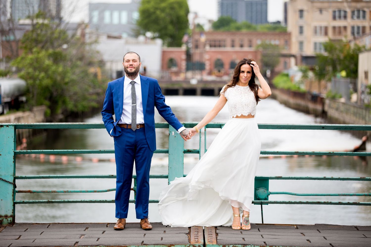 Couple in wedding attire poses stylishly on waterfront pier with urban backdrop.