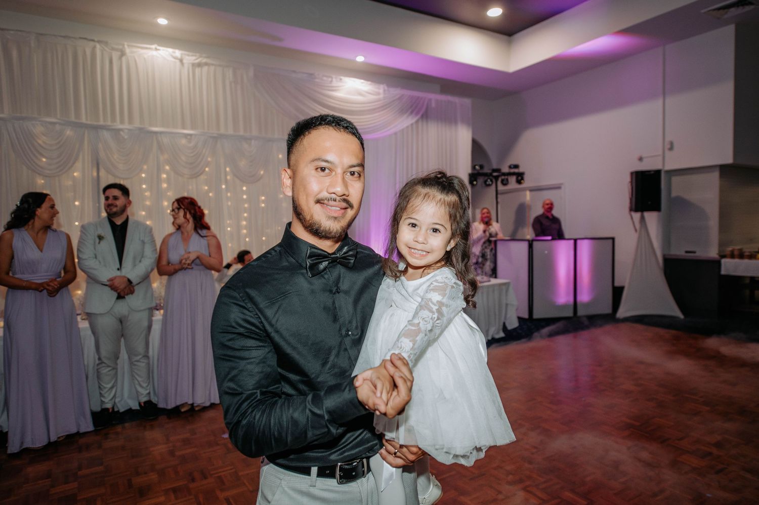 Parent dances with small child at wedding reception with purple lighting and guests in background.