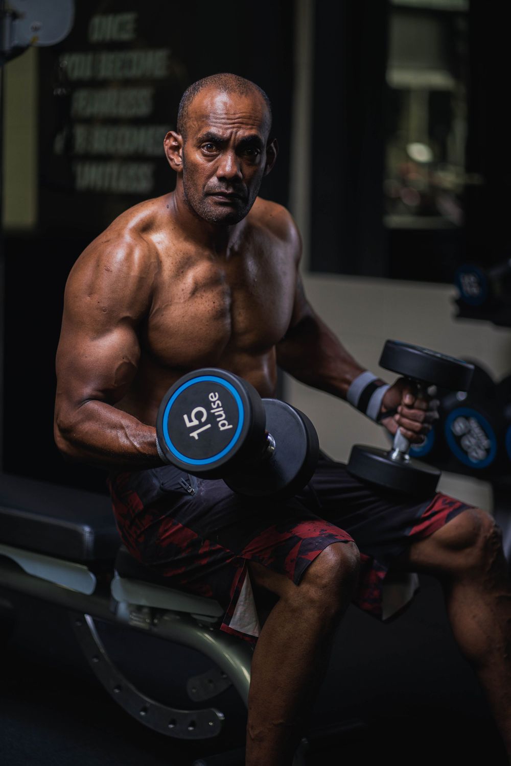 Athlete performs dumbbell exercise on bench showing muscular definition in dim gym lighting.