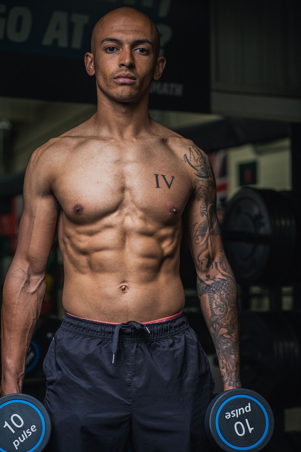 Muscular physique displayed during gym workout with blue dumbbells and visible tattoos.
