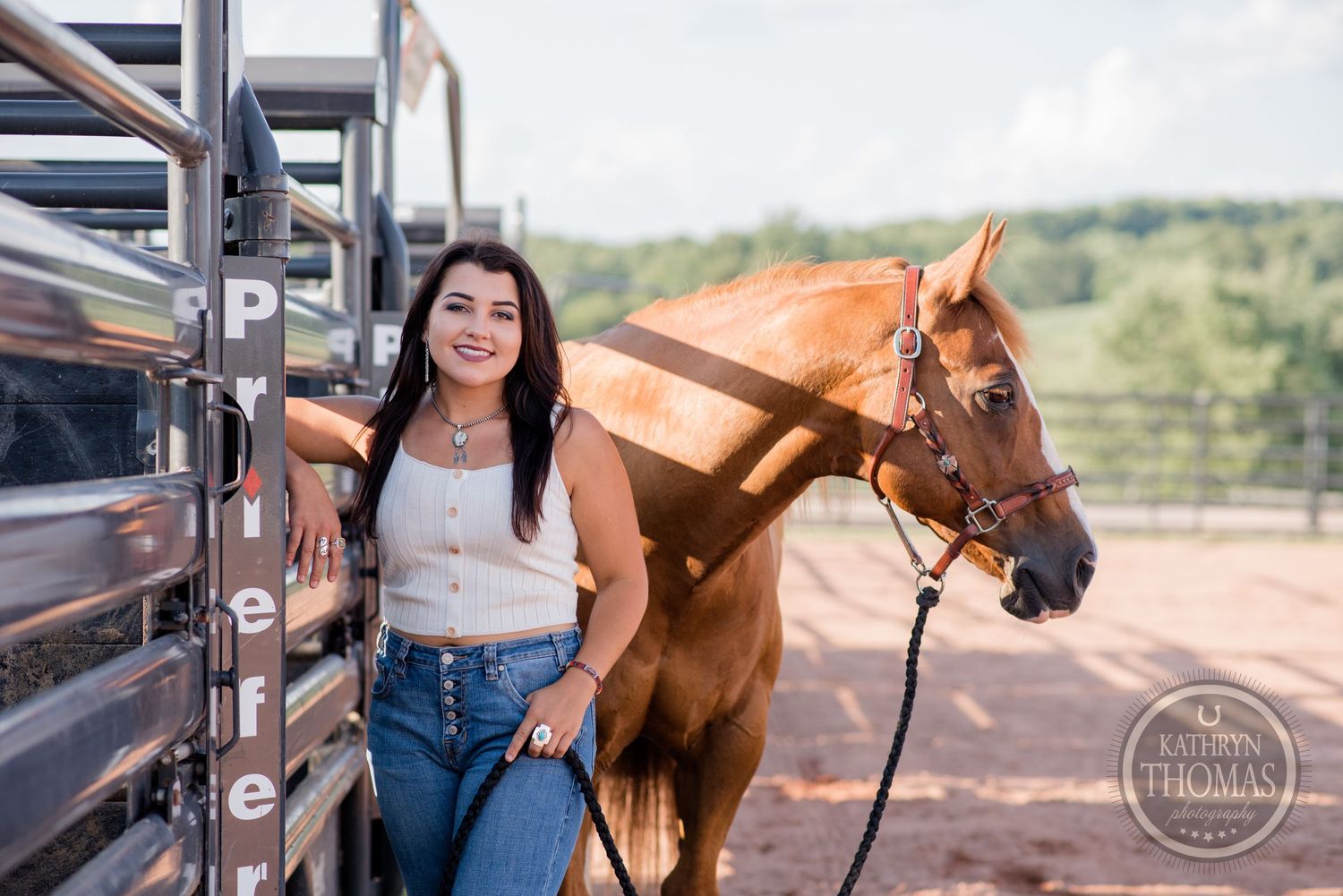 A chestnut horse stands beside metal horse trailer gates outdoors.