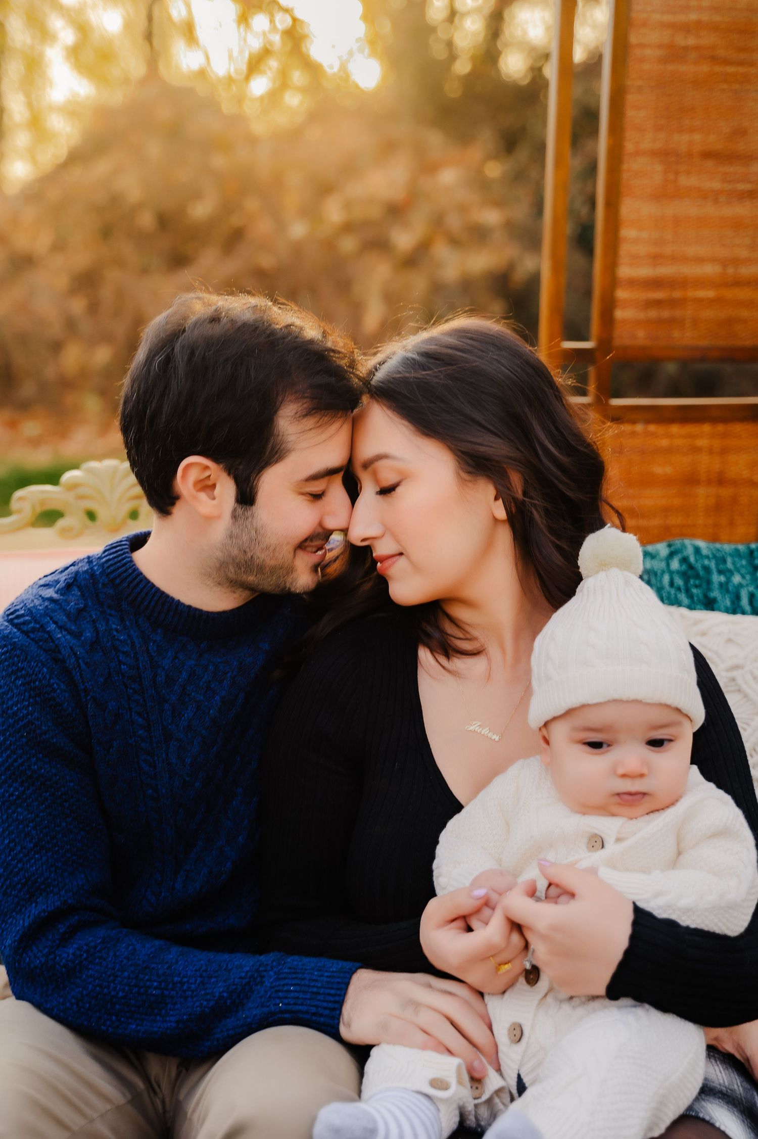 Young couple sitting with foreheads touching and young baby on their lap on Ft Wadsworth in Staten Island, NY
