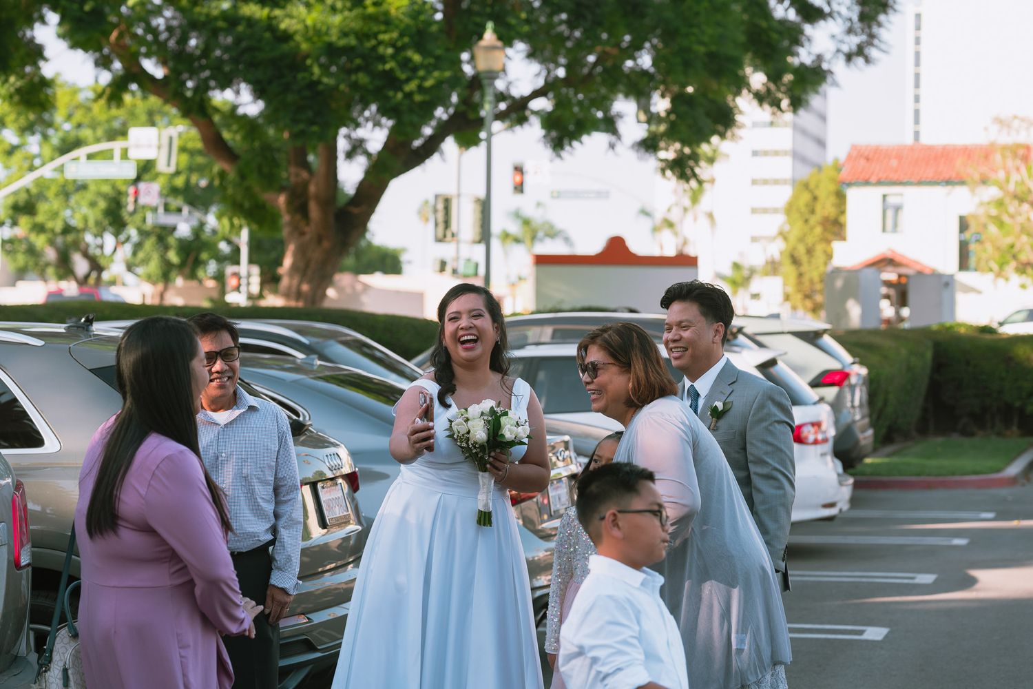 Bride in white gown laughing with wedding guests in sunny parking lot.