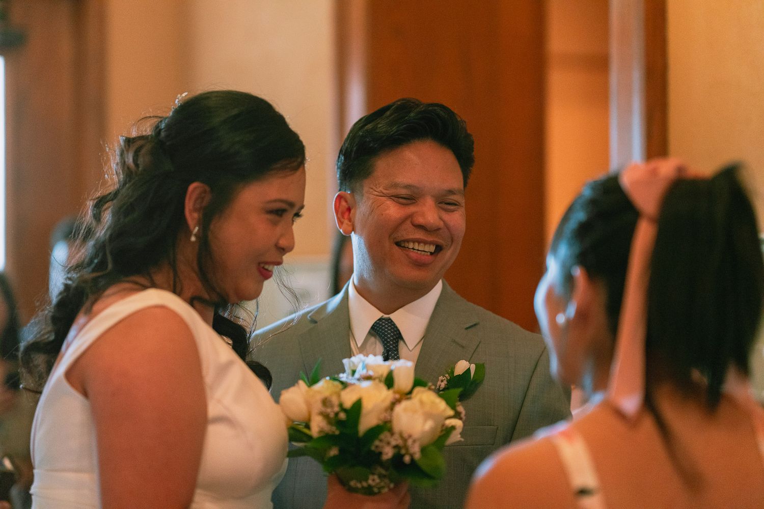 Bride and groom smiling together indoors holding white rose bouquet at wedding.