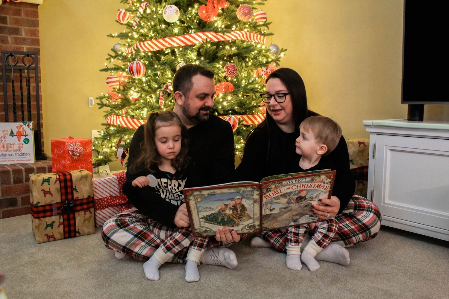 Family reads a book together in matching pajamas by a decorated Christmas tree surrounded by wrapped presents.