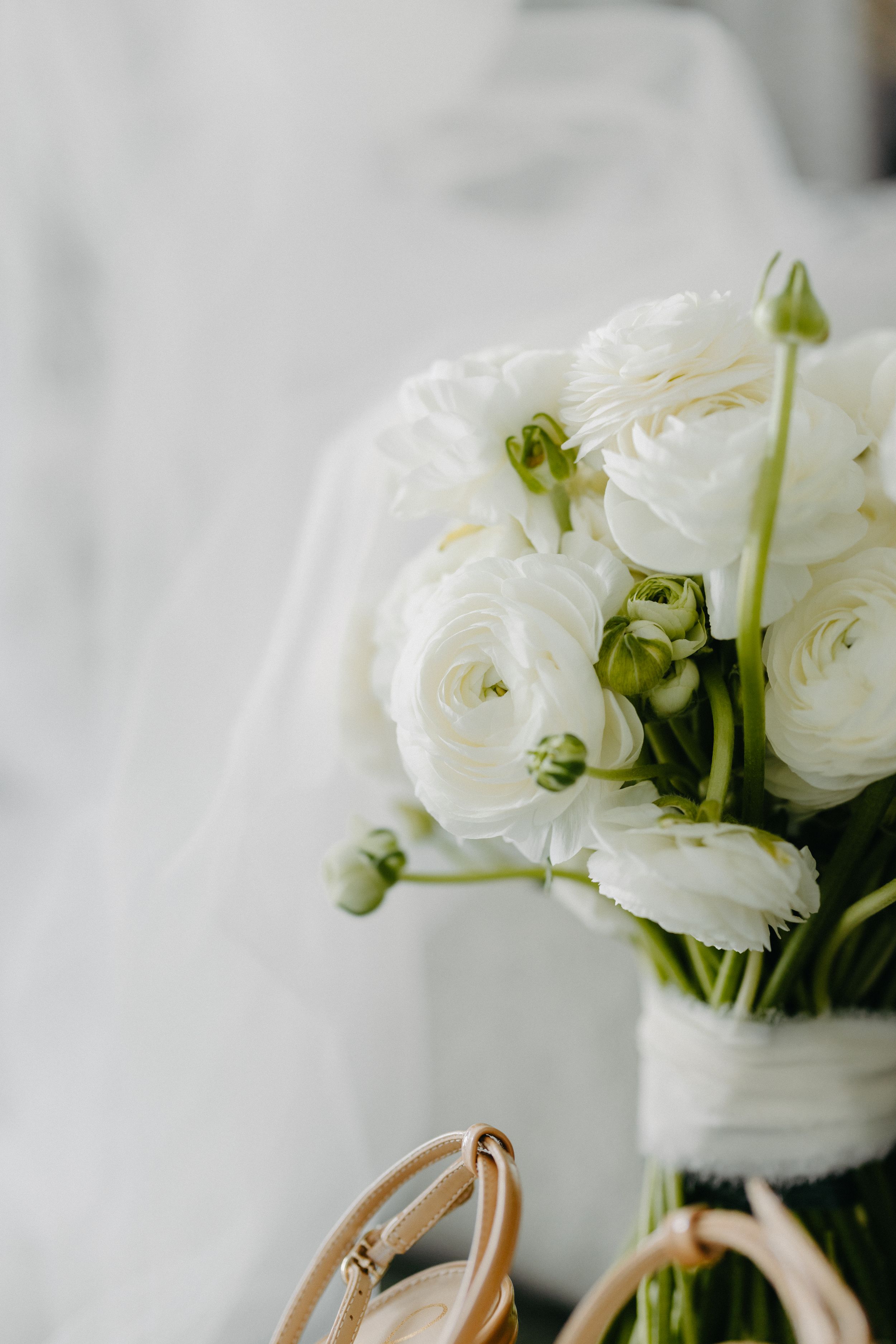 White ranunculus bouquet with green buds alongside bridal shoes against light background.