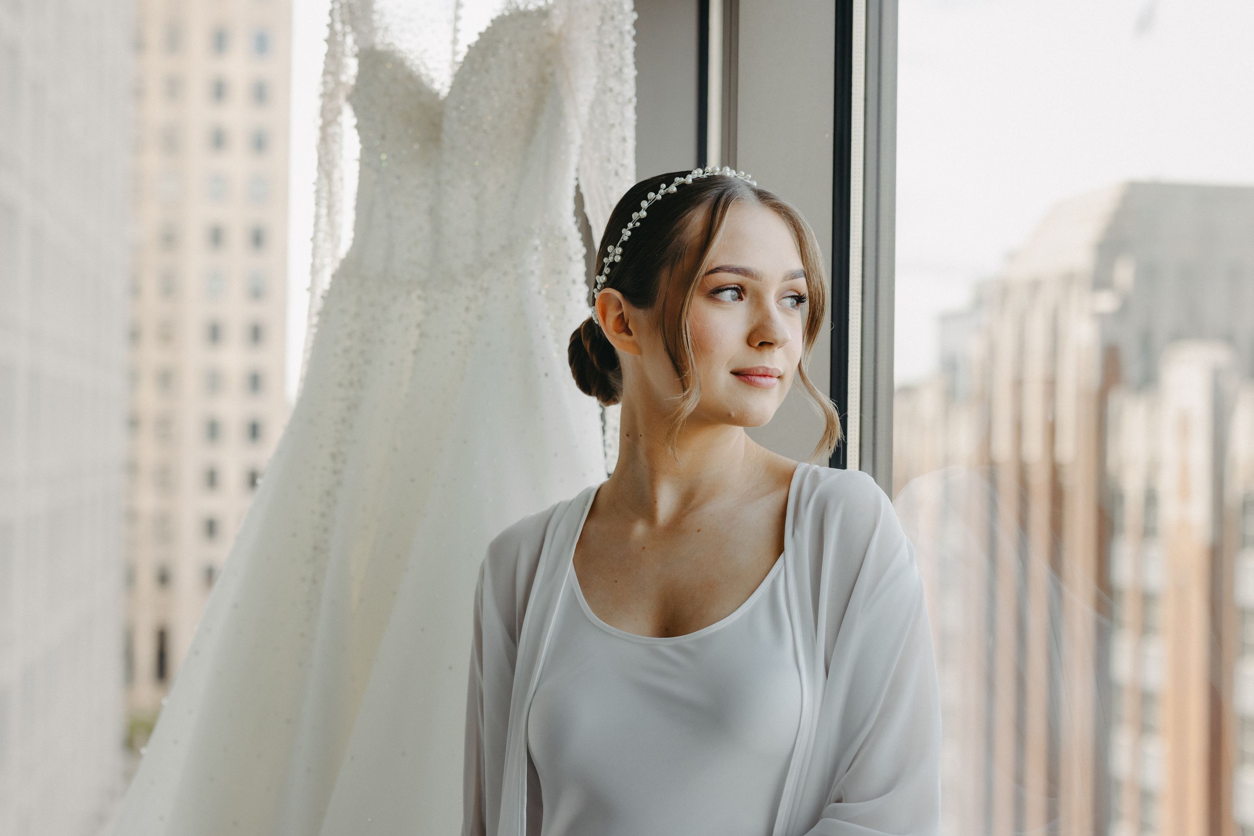 Bride in white robe and headphones looking out window with urban background.
