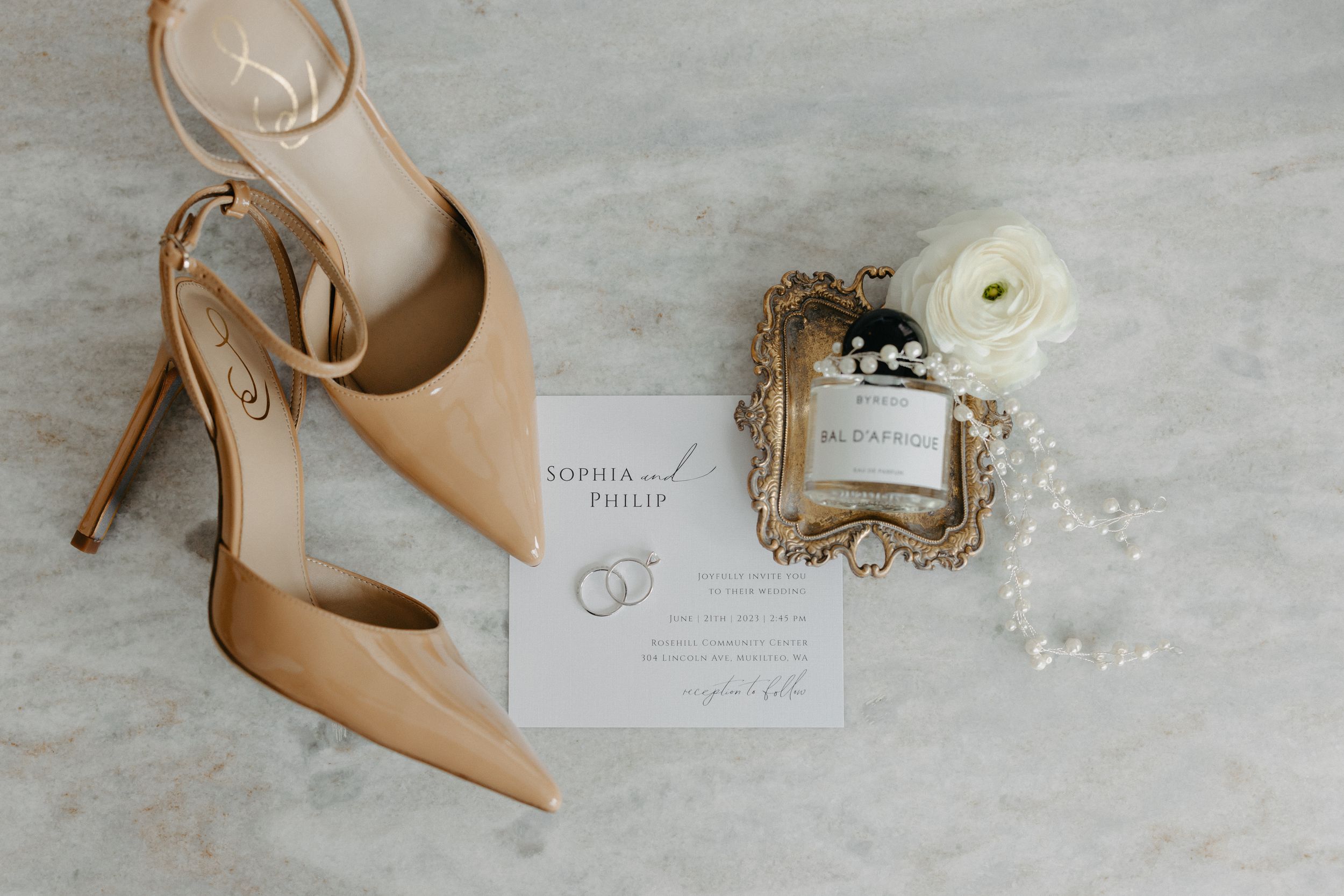 Wedding flat lay with nude heels, perfume bottle, and invitation card on marble surface.