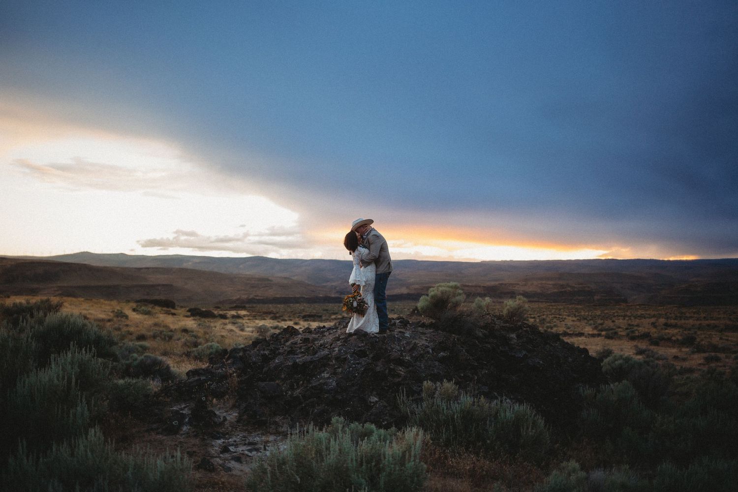 A groom dips back and kisses his bride on a giant rock with a beautiful sunset behind them in the desert