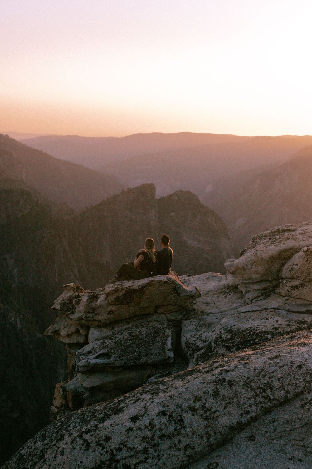 Two people stand on rocky cliffs overlooking misty mountains at sunset in a scenic wilderness landscape.