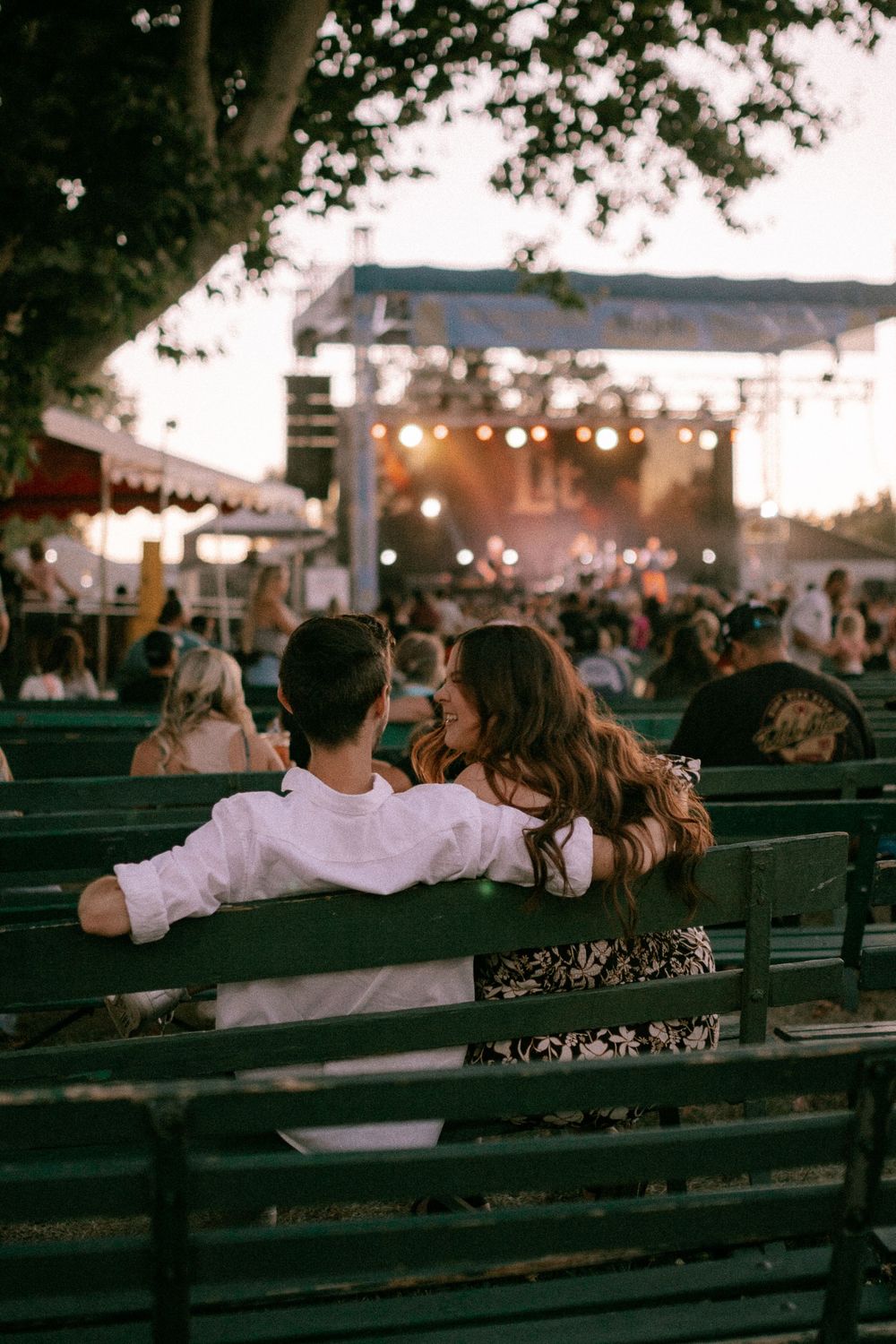 Couple enjoying an outdoor concert while sitting on wooden benches at dusk.