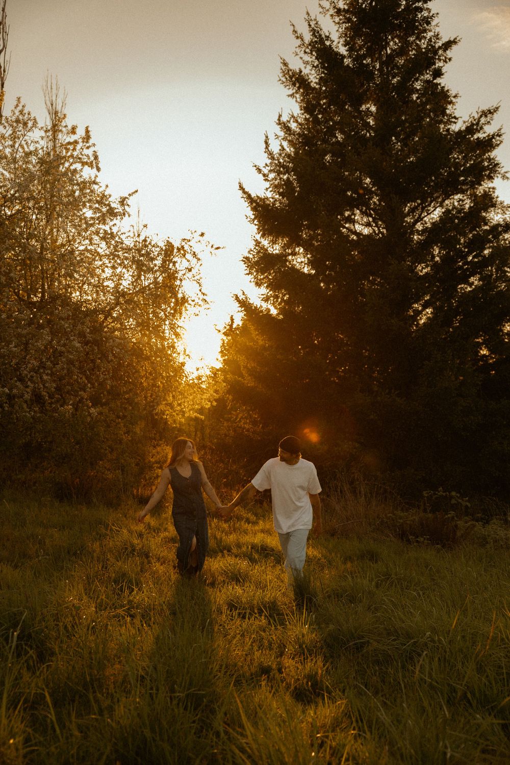 Couple holding hands walking through a sunlit meadow surrounded by trees during golden hour sunset.