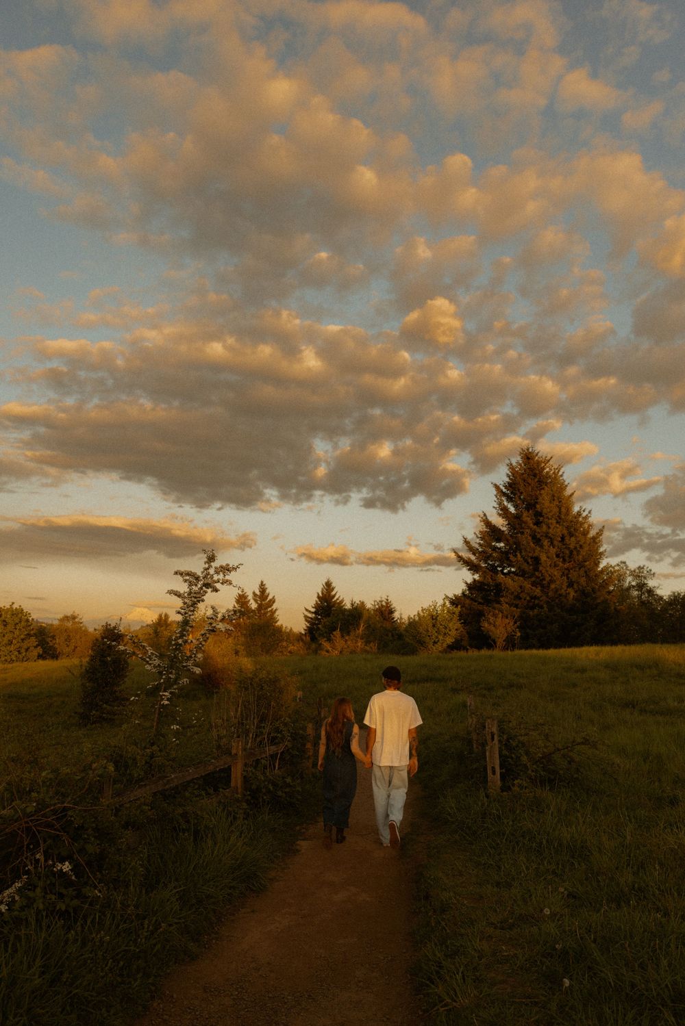 Couple holding hands walking on a dirt path through green fields at golden sunset with dramatic clouds overhead.