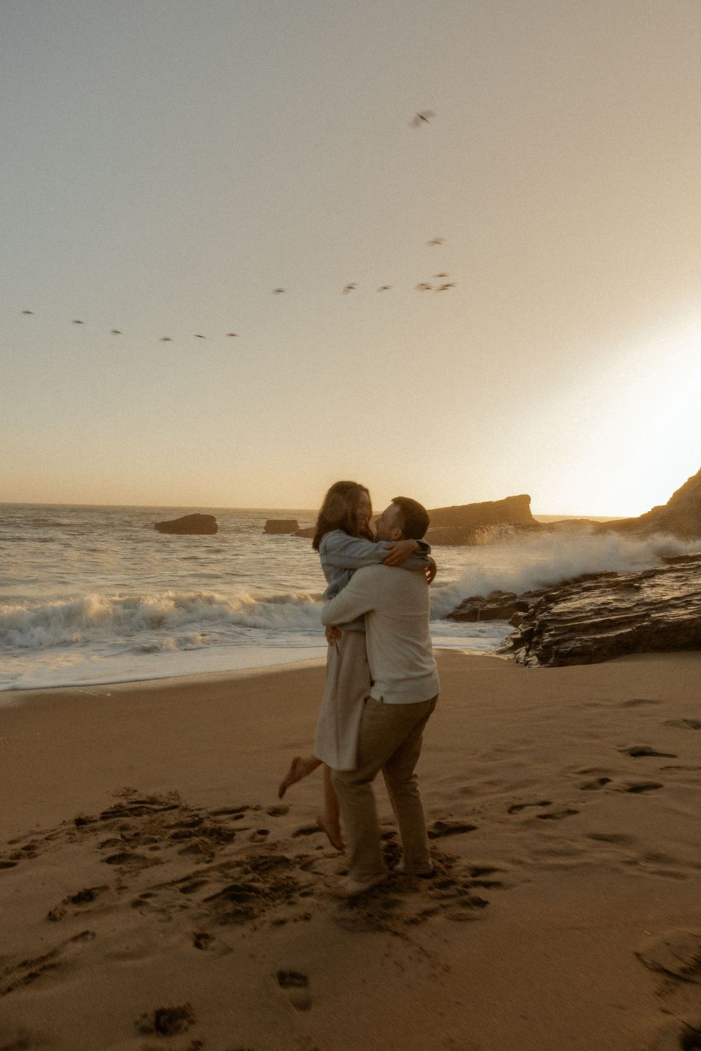 Couple embracing on a sandy beach at golden sunset with ocean waves and birds flying overhead.
