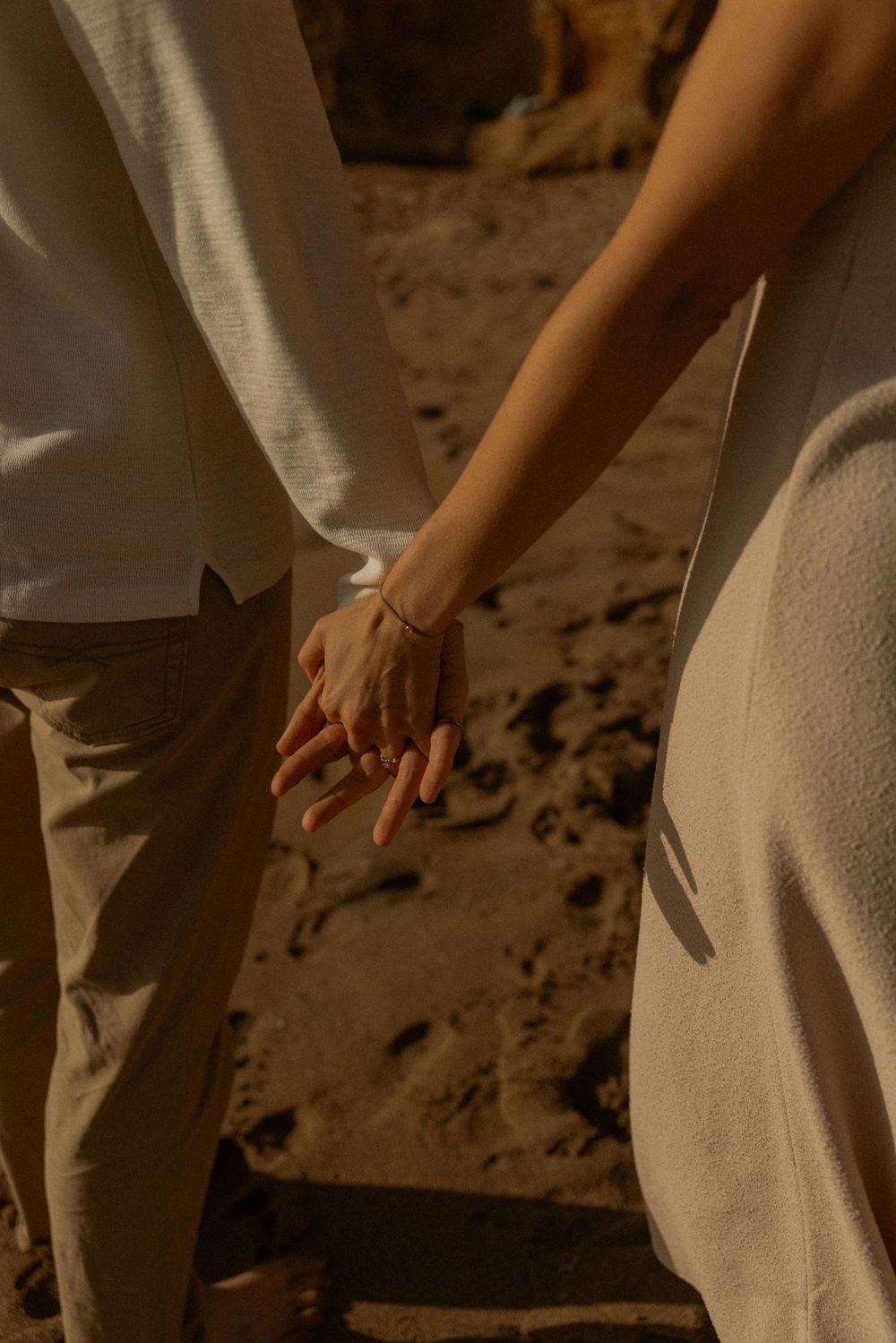Couple holding hands on sandy beach, wearing neutral tones, with a gold bracelet visible, captured in warm golden light.