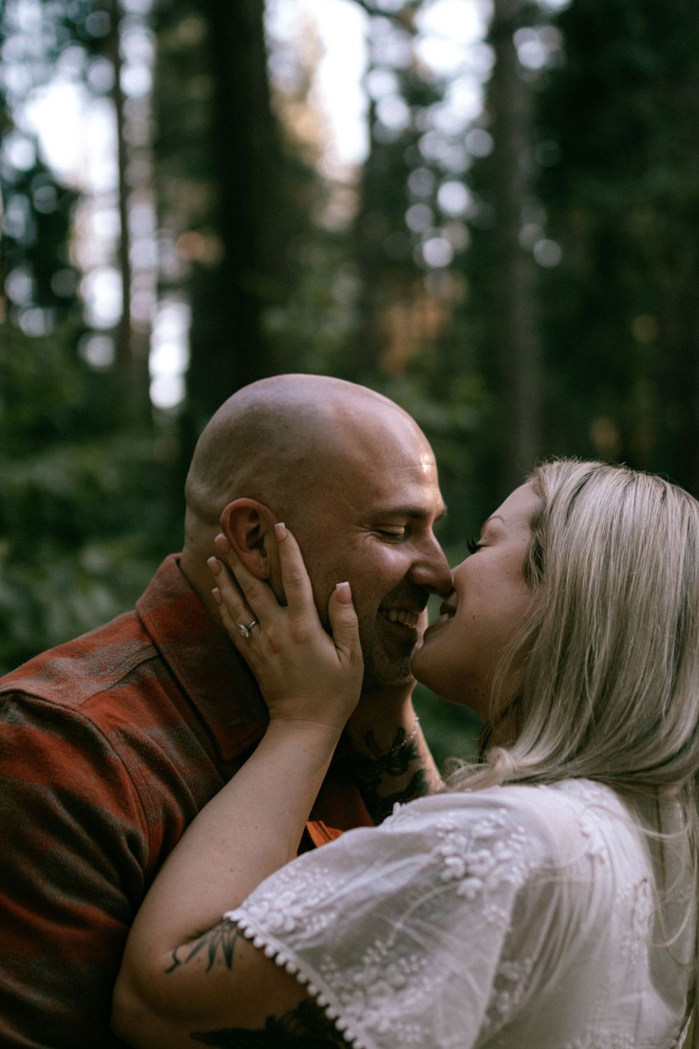 A couple shares an intimate kiss in the woods wearing rustic clothing against a blurred forest backdrop.