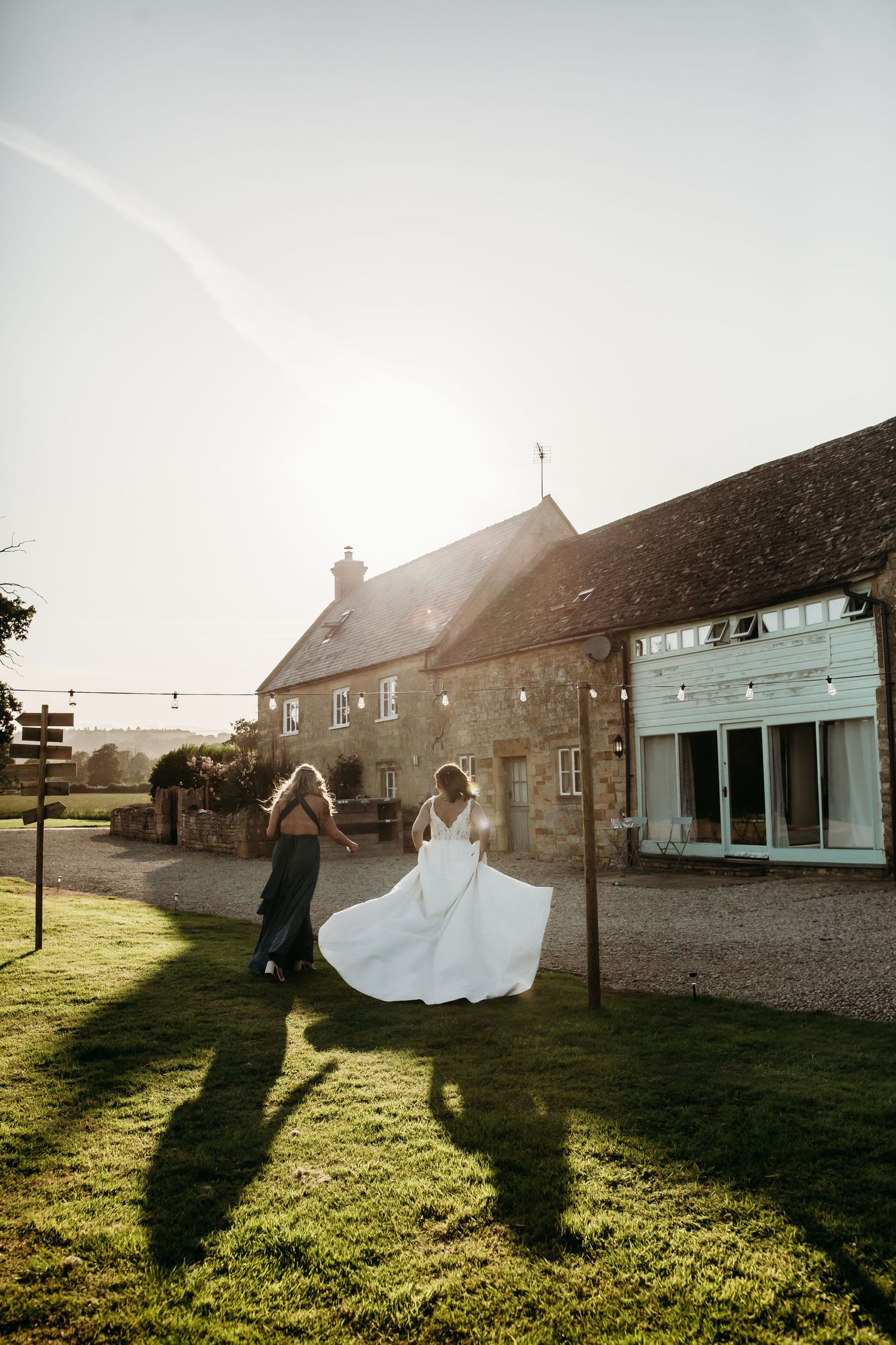 A couple in wedding attire twirls in golden sunlight on a grassy lawn next to stone buildings.