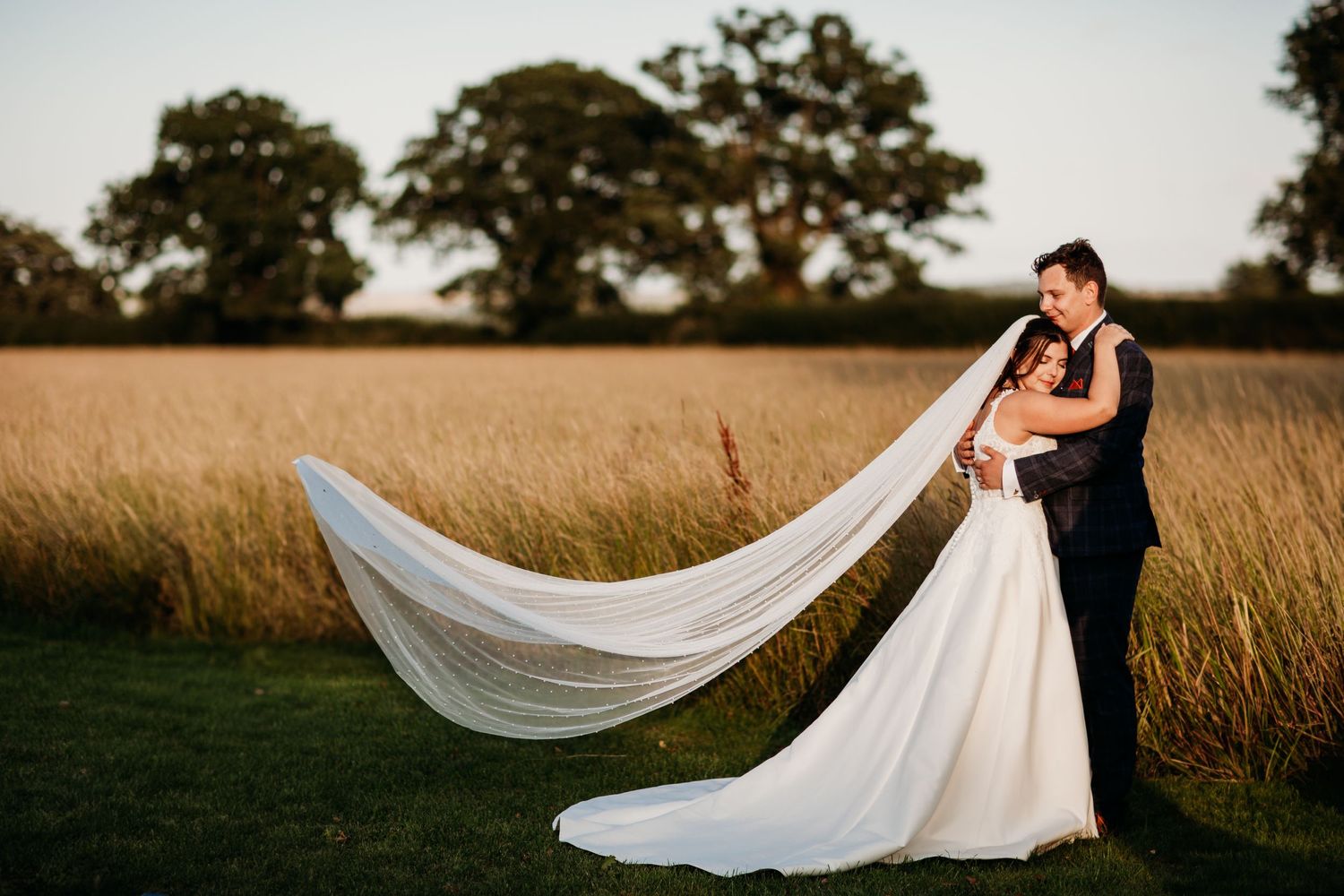 Couple in wedding attire pose in a golden field with flowing veil catching the wind.