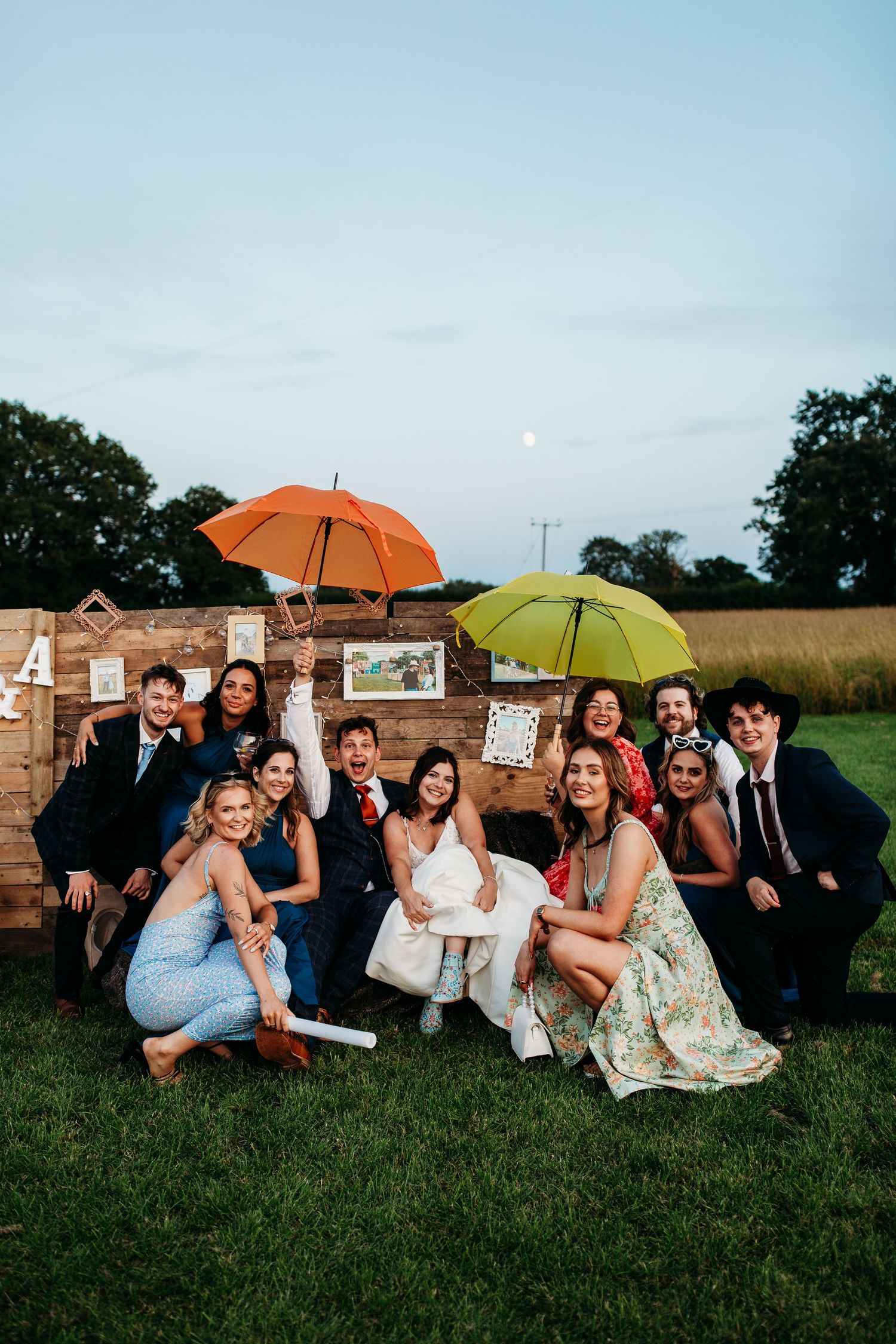 Wedding party poses with colorful umbrellas against a rustic stone wall at dusk.