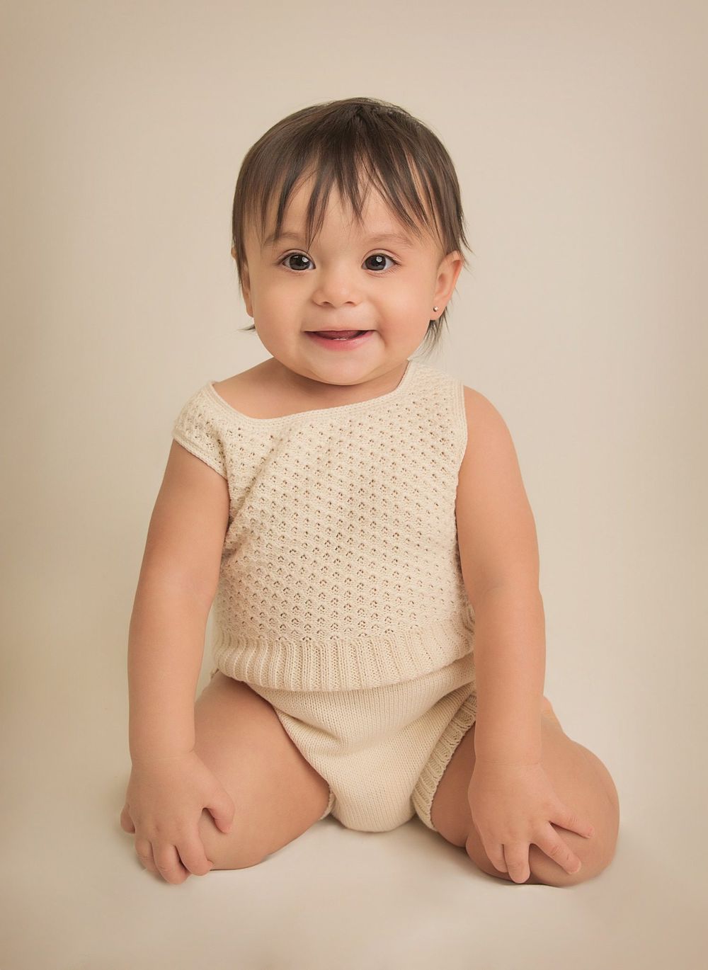 Baby wearing cream knit outfit sits on neutral background in studio setting.