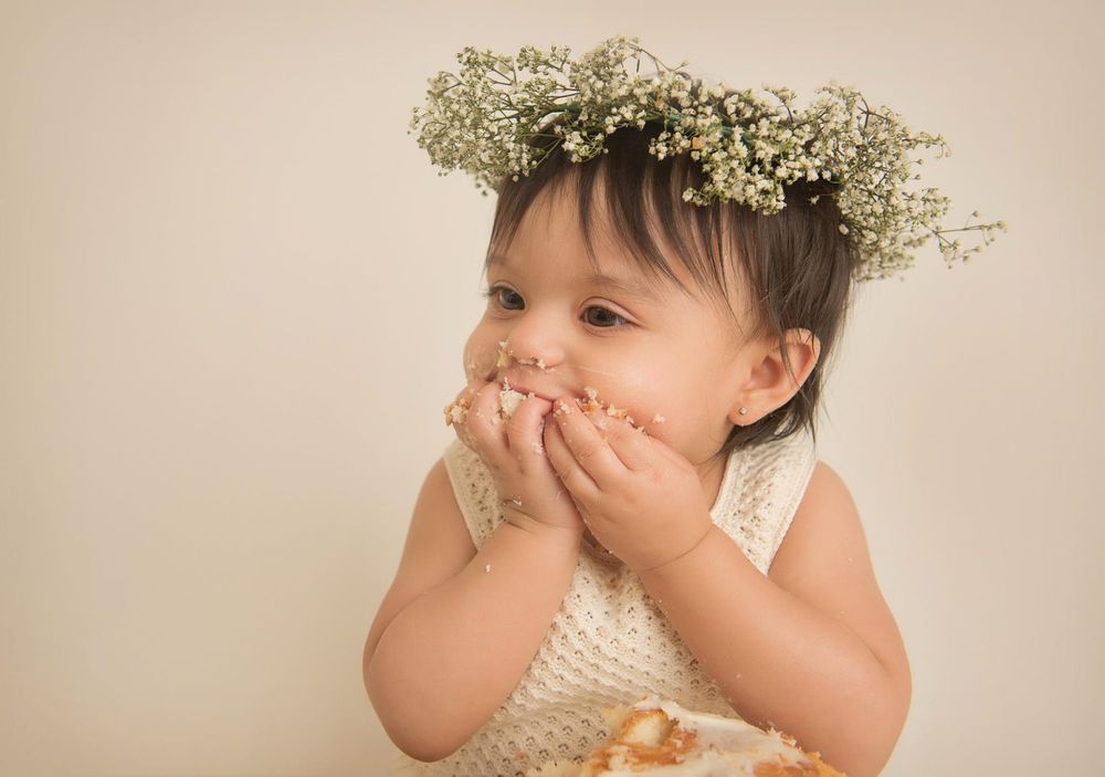 Baby wearing floral crown and cream outfit giggles with hands covering mouth.