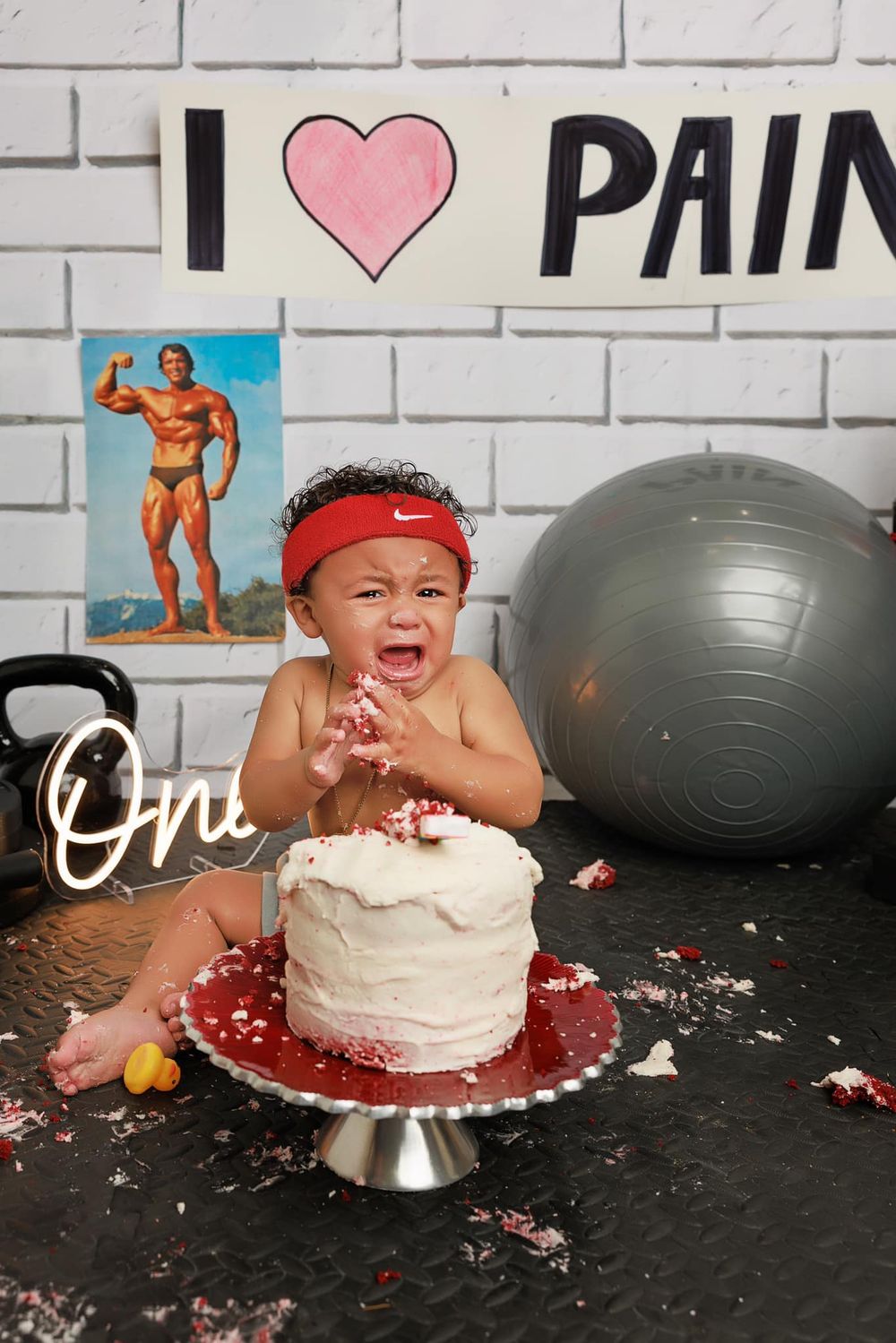 Baby wearing red headband enjoys first birthday cake smash against gym-themed backdrop.