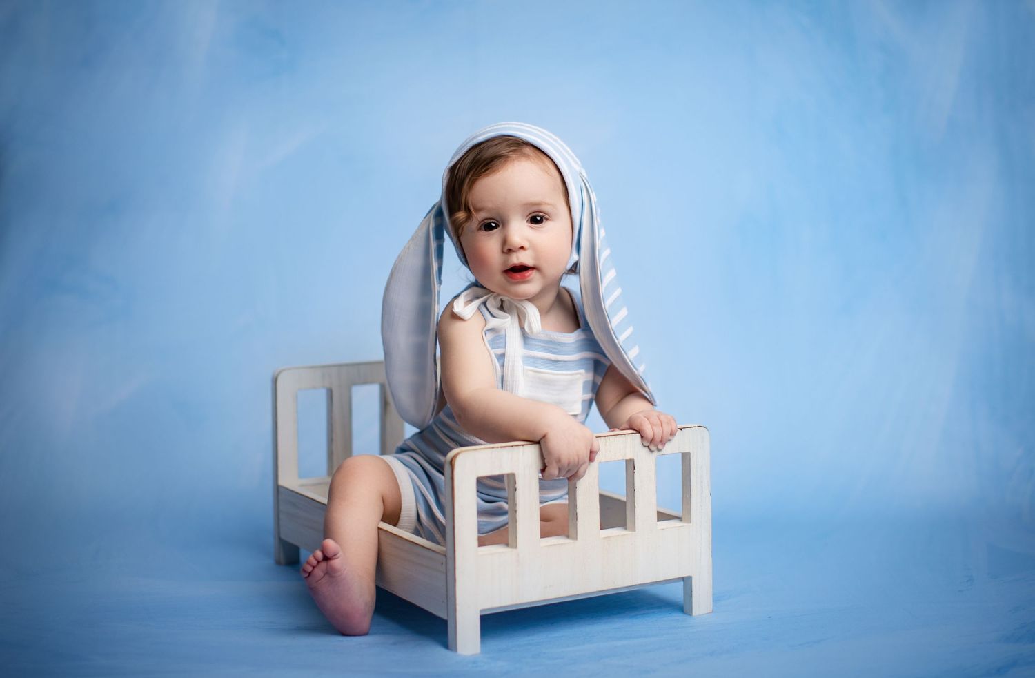 Small child in white outfit sitting in miniature bed against blue background.