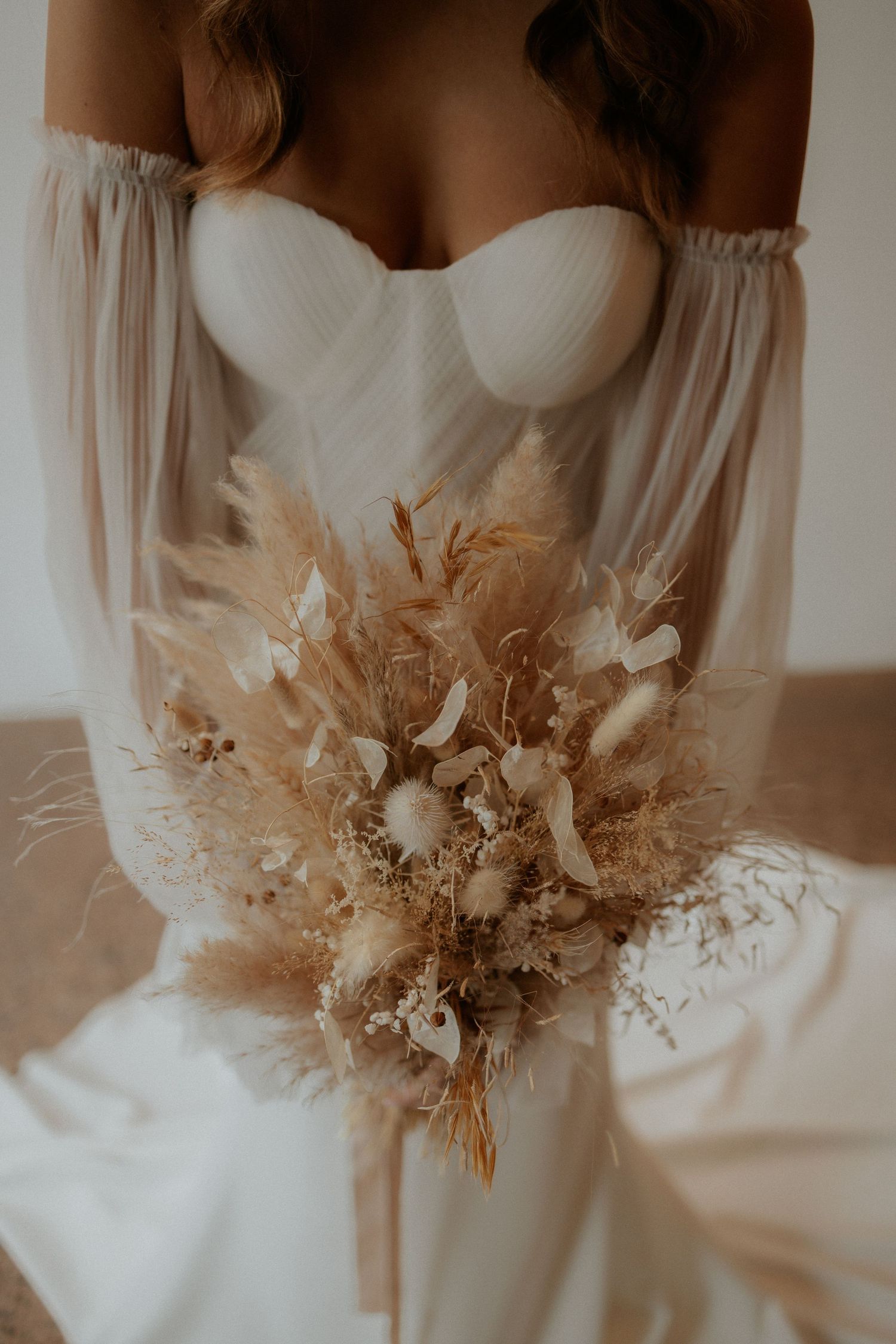 A romantic white wedding dress with an off-shoulder design and dried pampas grass bouquet in warm neutral tones.