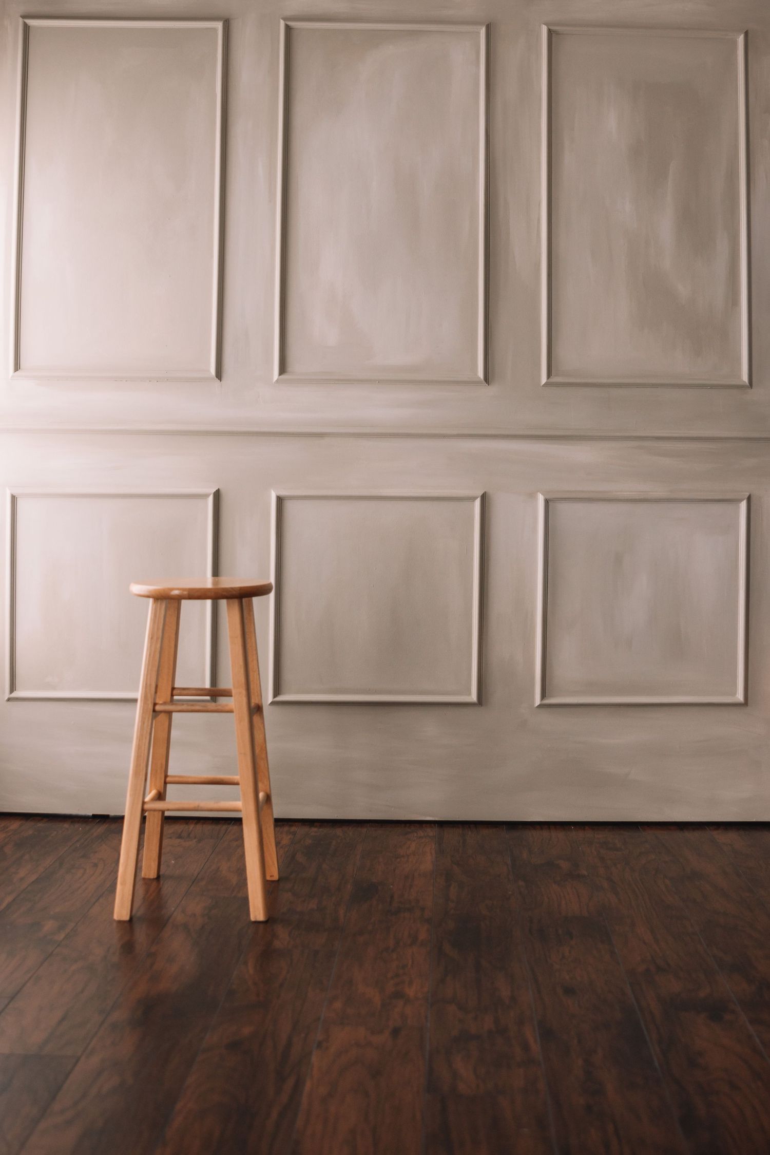 Tall wooden barstool against beige paneled wall with dark hardwood floors.