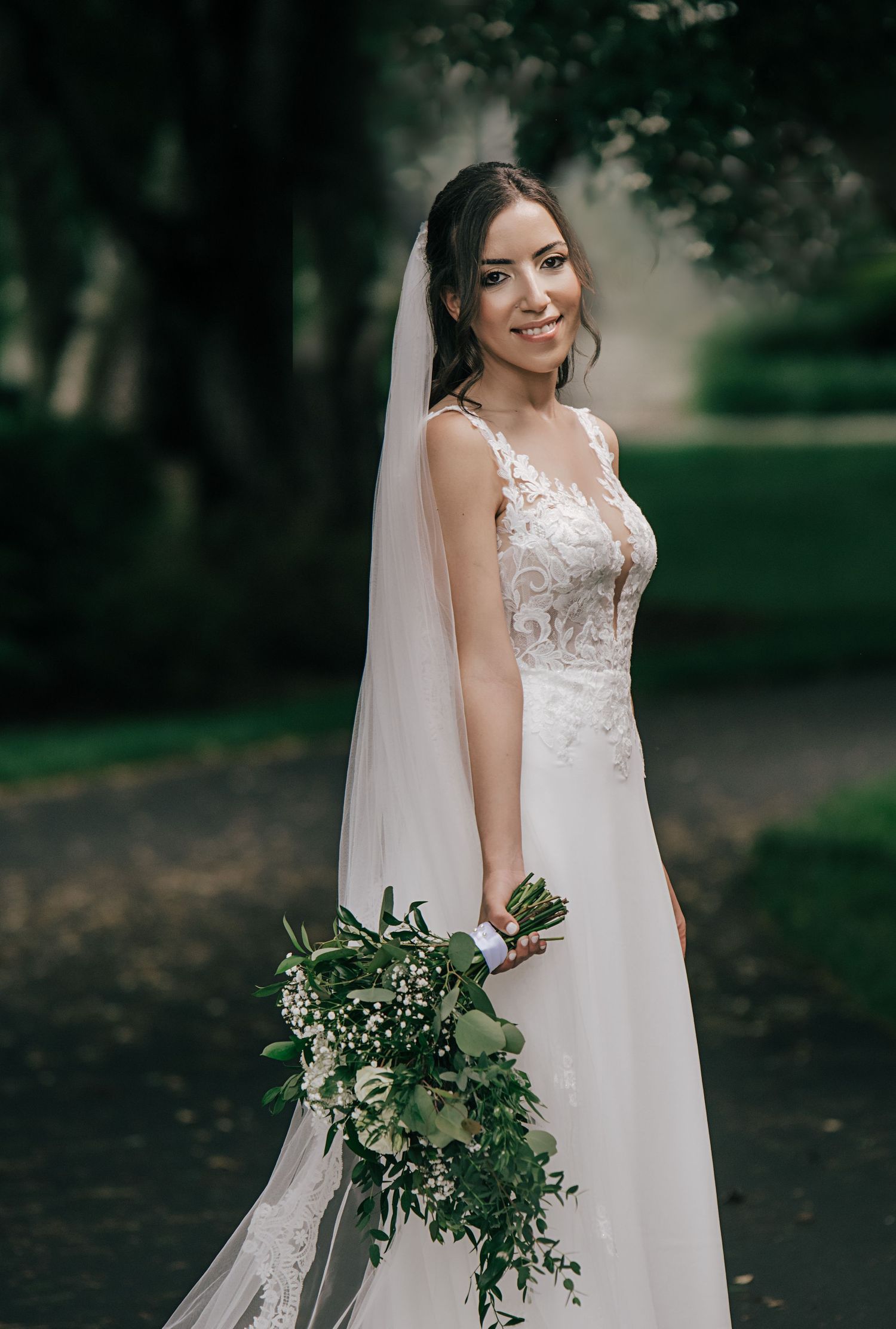 Bride in lace detailed gown holds elegant greenery bouquet in garden setting.