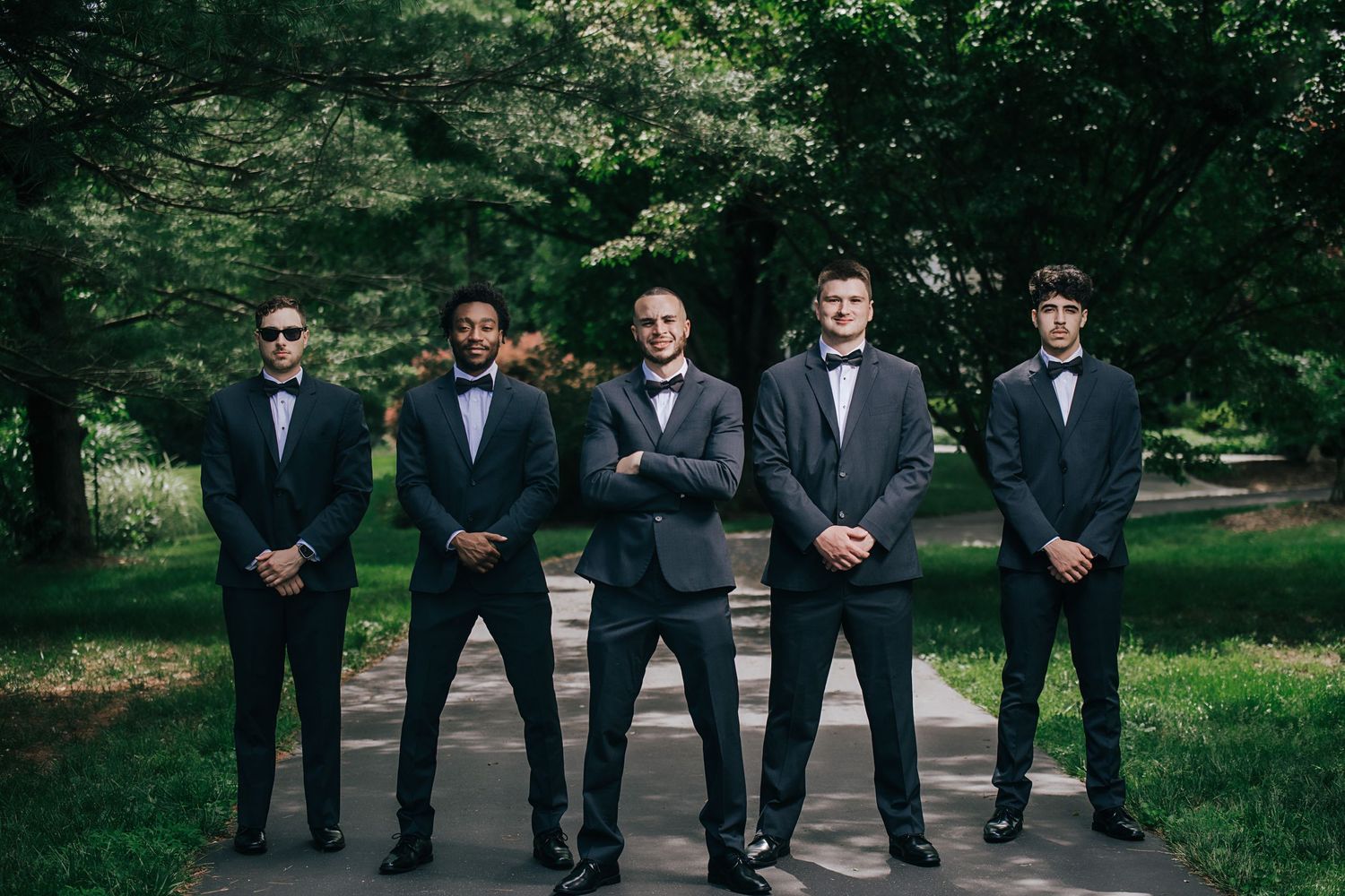 Groomsmen in black tuxedos pose together on garden path.