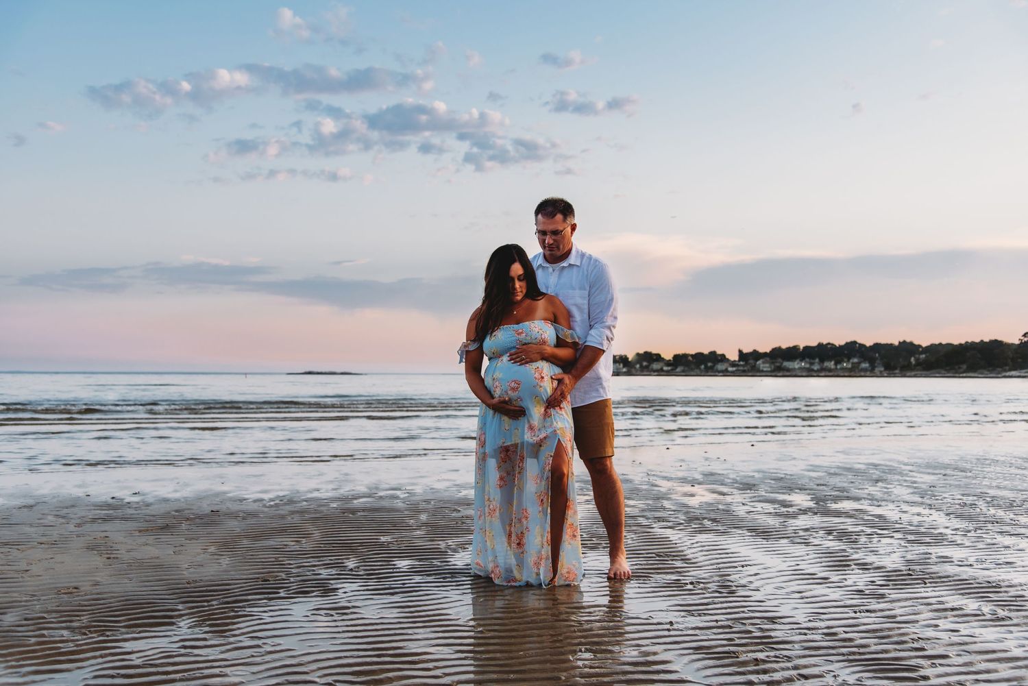Couple standing in shallow ocean water at sunset during maternity photoshoot.