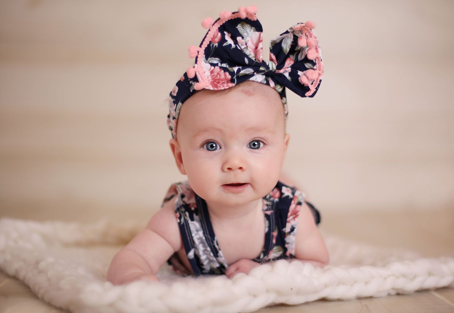 Baby wearing floral outfit and bow headband lying on soft pink blanket.