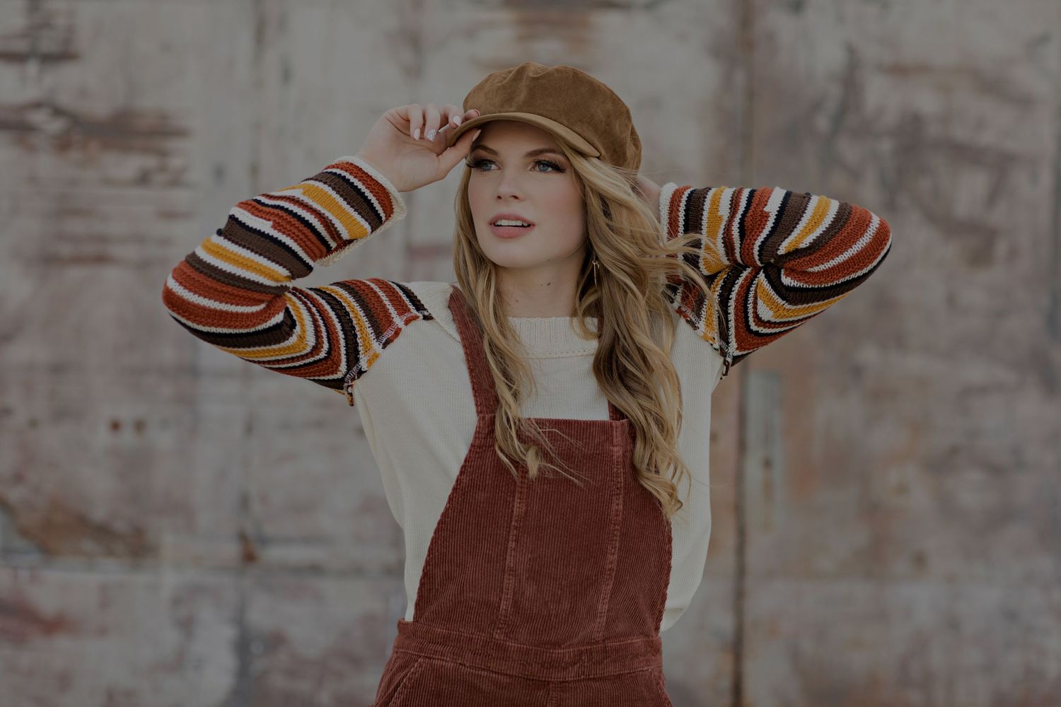 Model wearing brown corduroy overalls and striped long sleeve top poses against concrete wall.