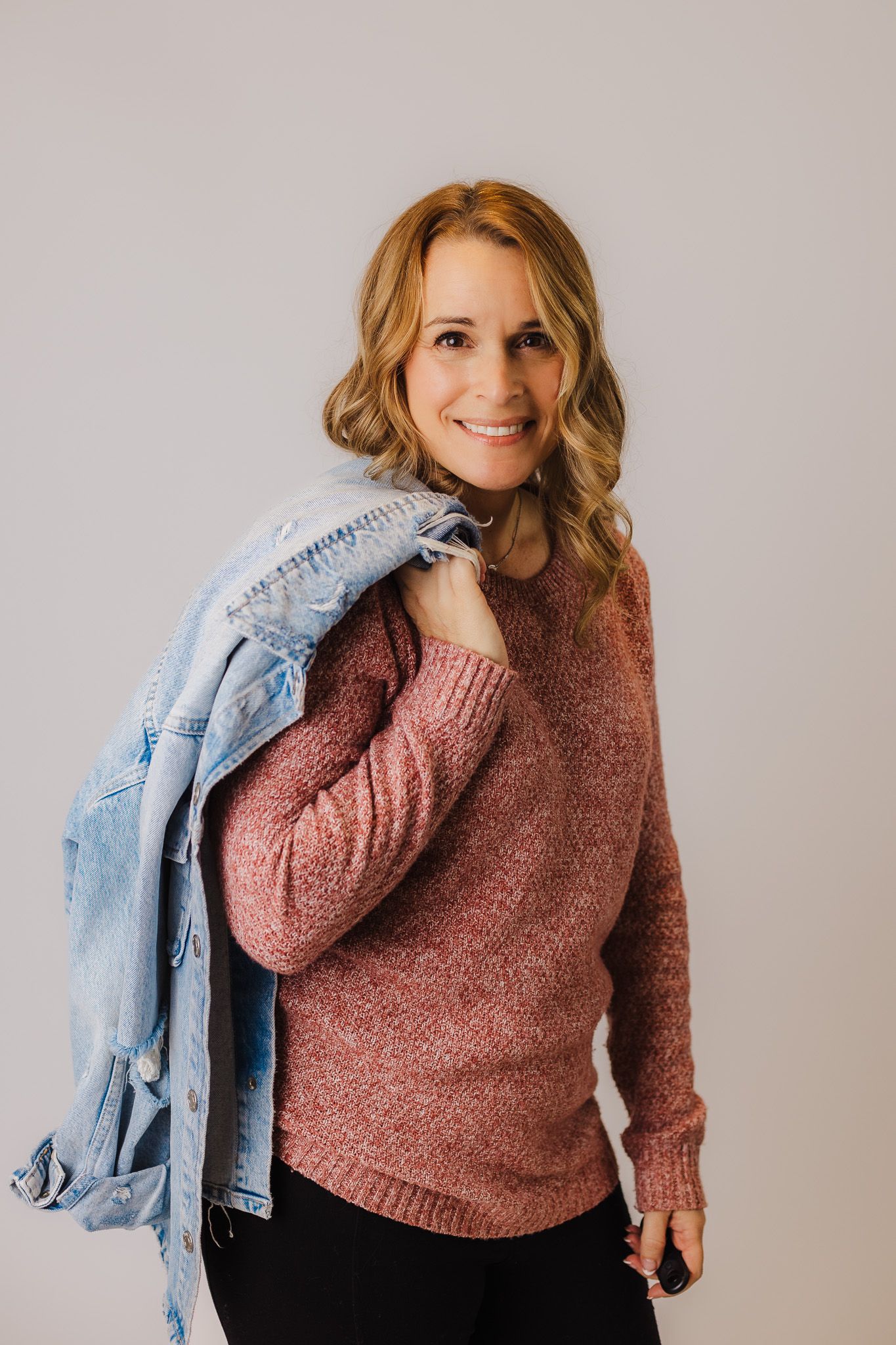 Person wearing pink sweater and denim jacket posing against neutral gray background.
