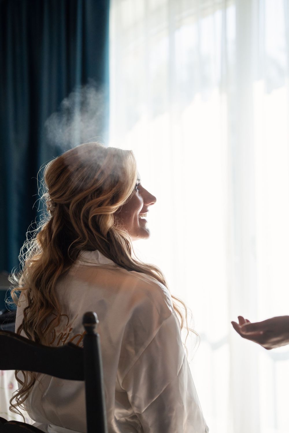 Silhouette of bridal preparation against backlit white curtains.