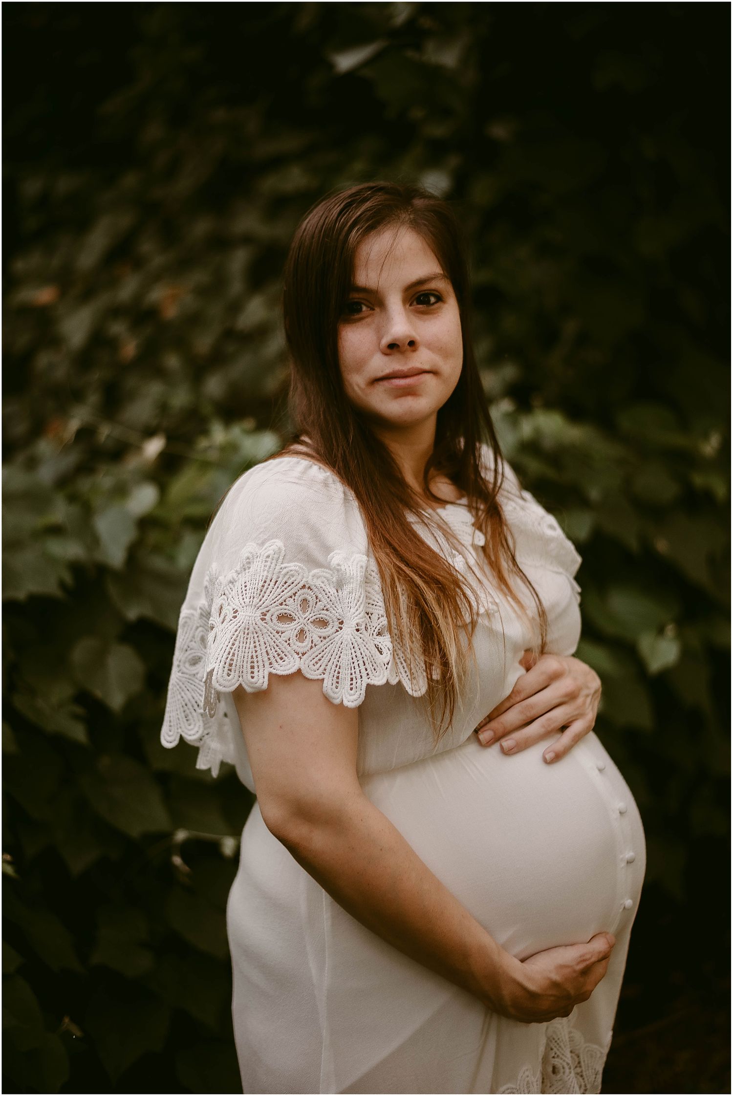 Expectant mother in white lace dress poses against green foliage background.